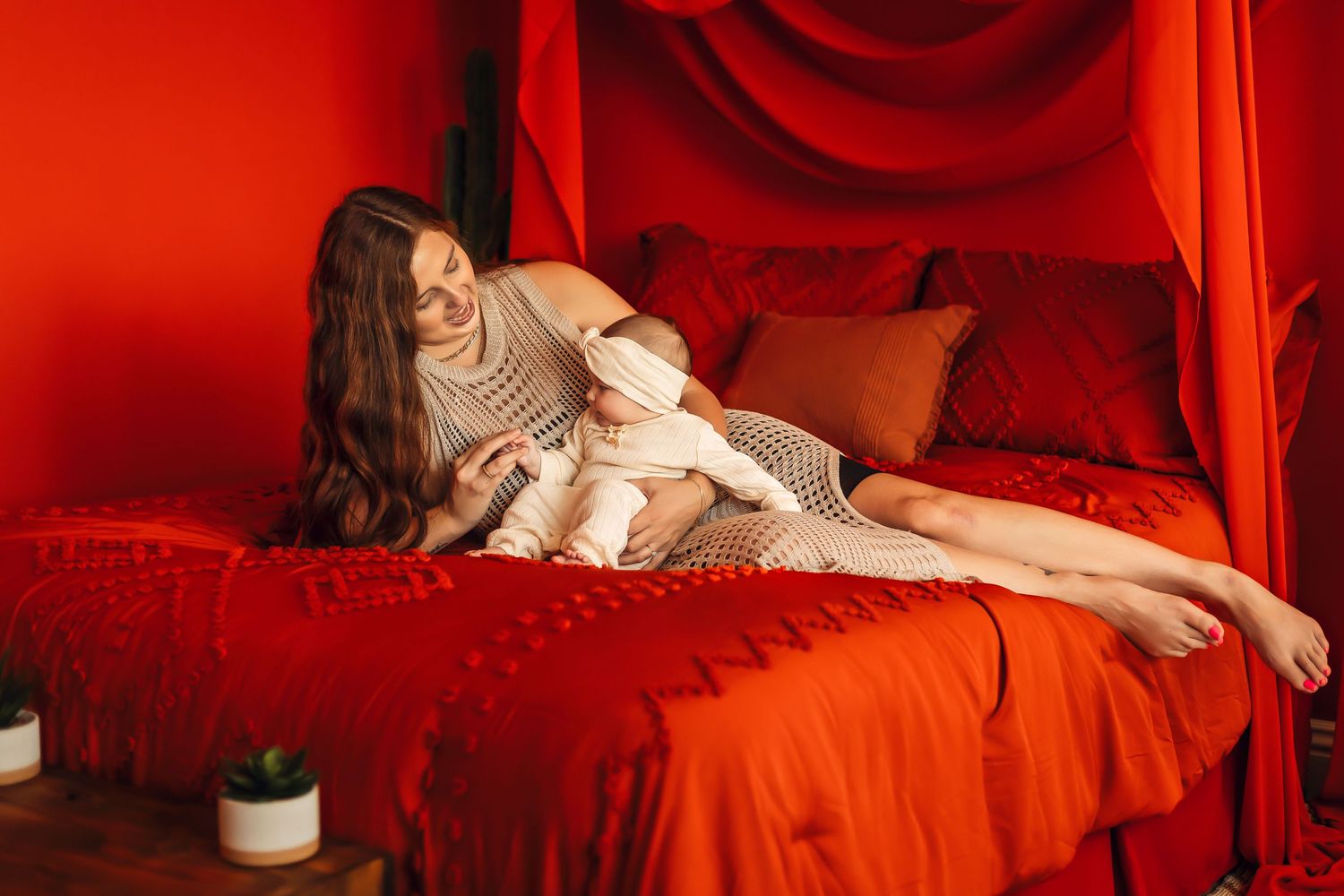 A parent and baby relax together on a red bed in a warmly lit room with minimalist decor.