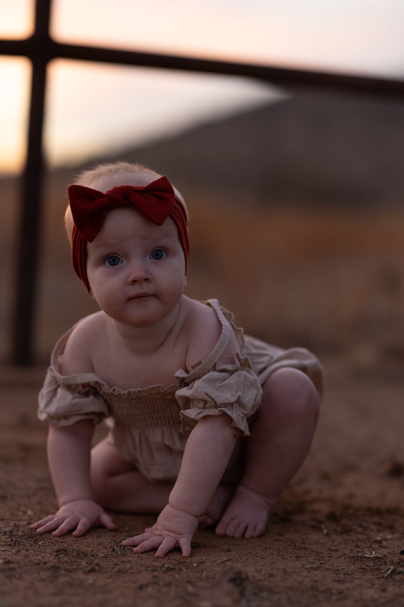 A baby in a beige dress and red bow headband crawls on dirt ground at sunset with window silhouette in background.