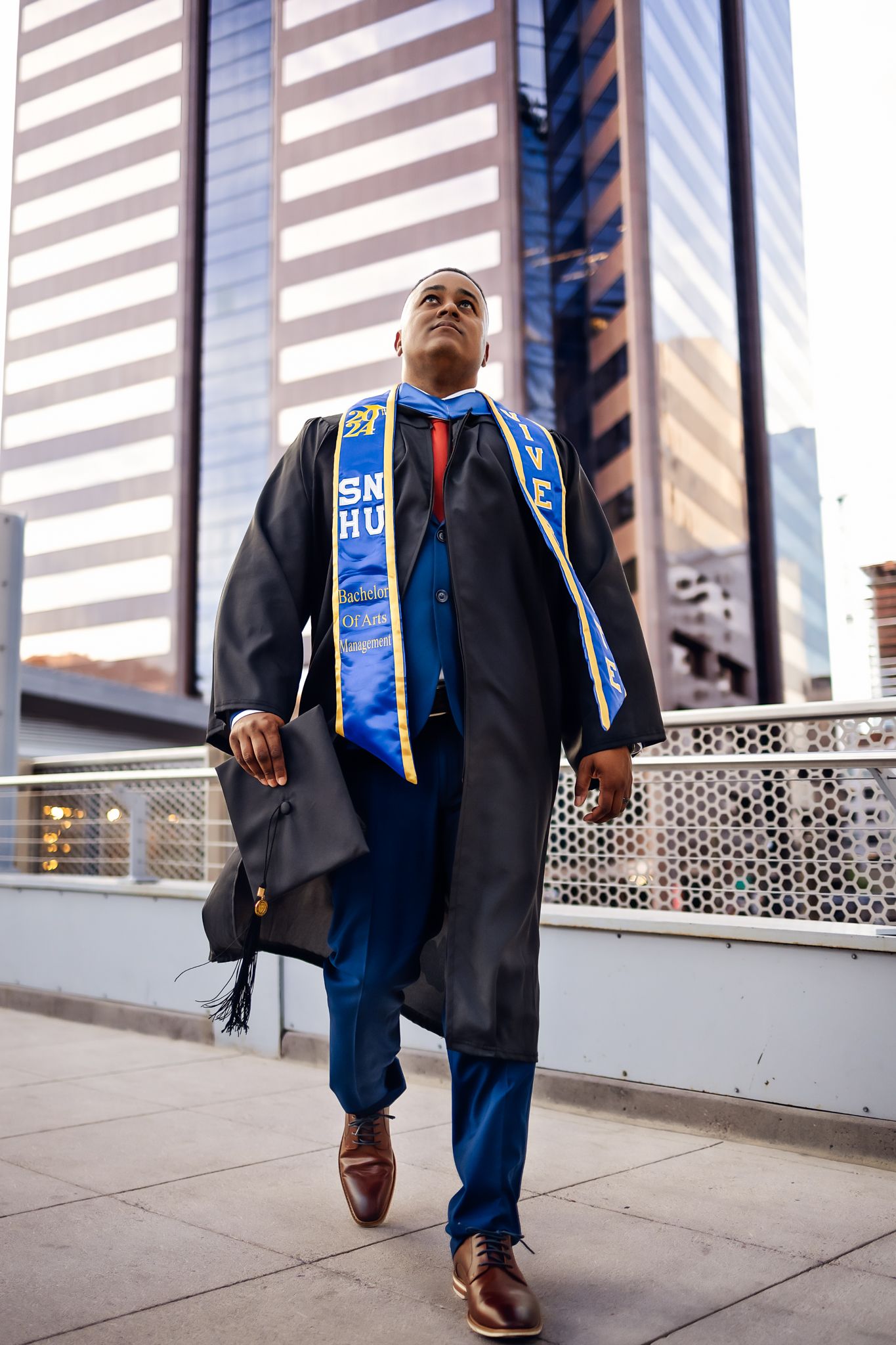 Graduate in cap and gown with honor cords poses confidently in front of modern office building in Downtown Phoenix.
