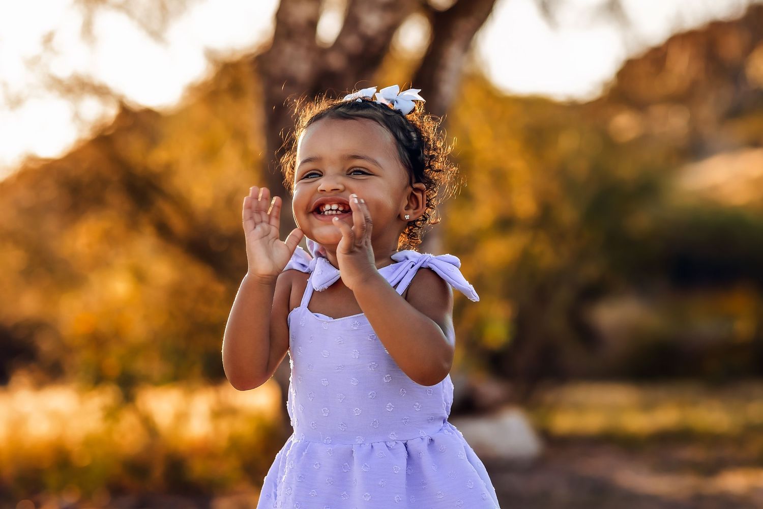 A joyful toddler in white dress laughs while playing in golden sunset light.