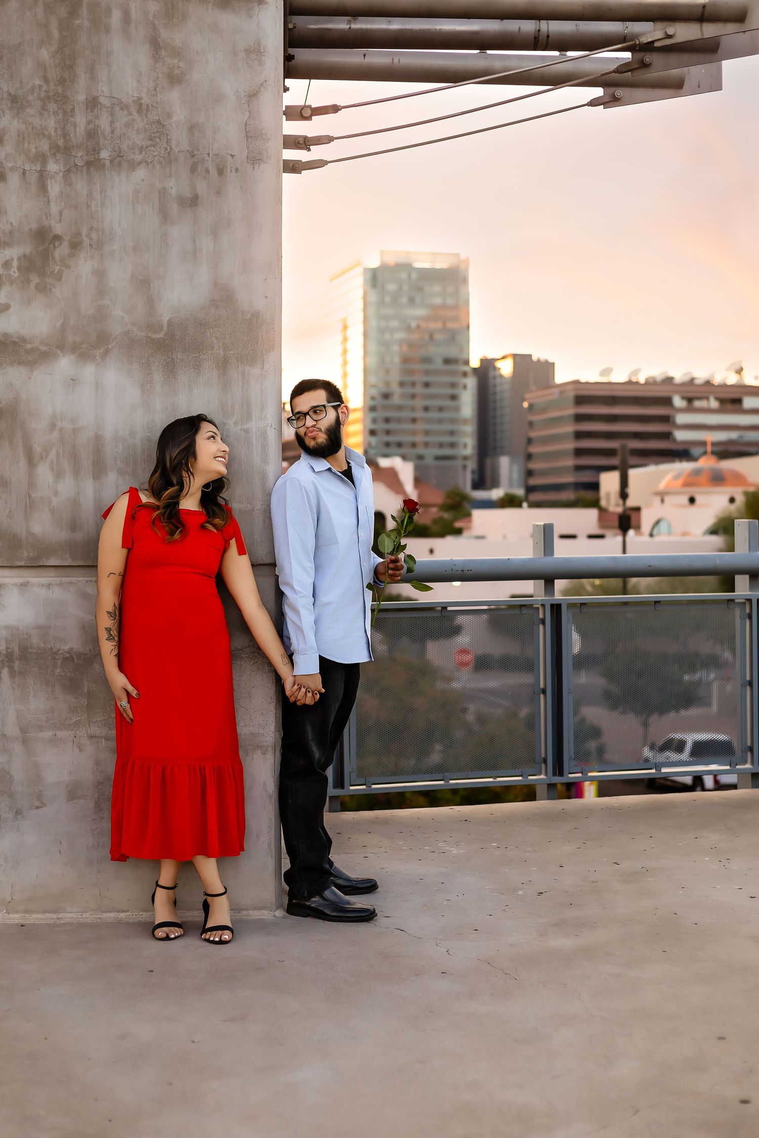 Couple in contrasting red dress and blue shirt share a playful moment on urban rooftop at sunset.