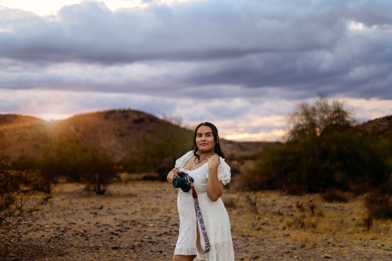 Photographer in white dress poses against desert landscape at sunset.