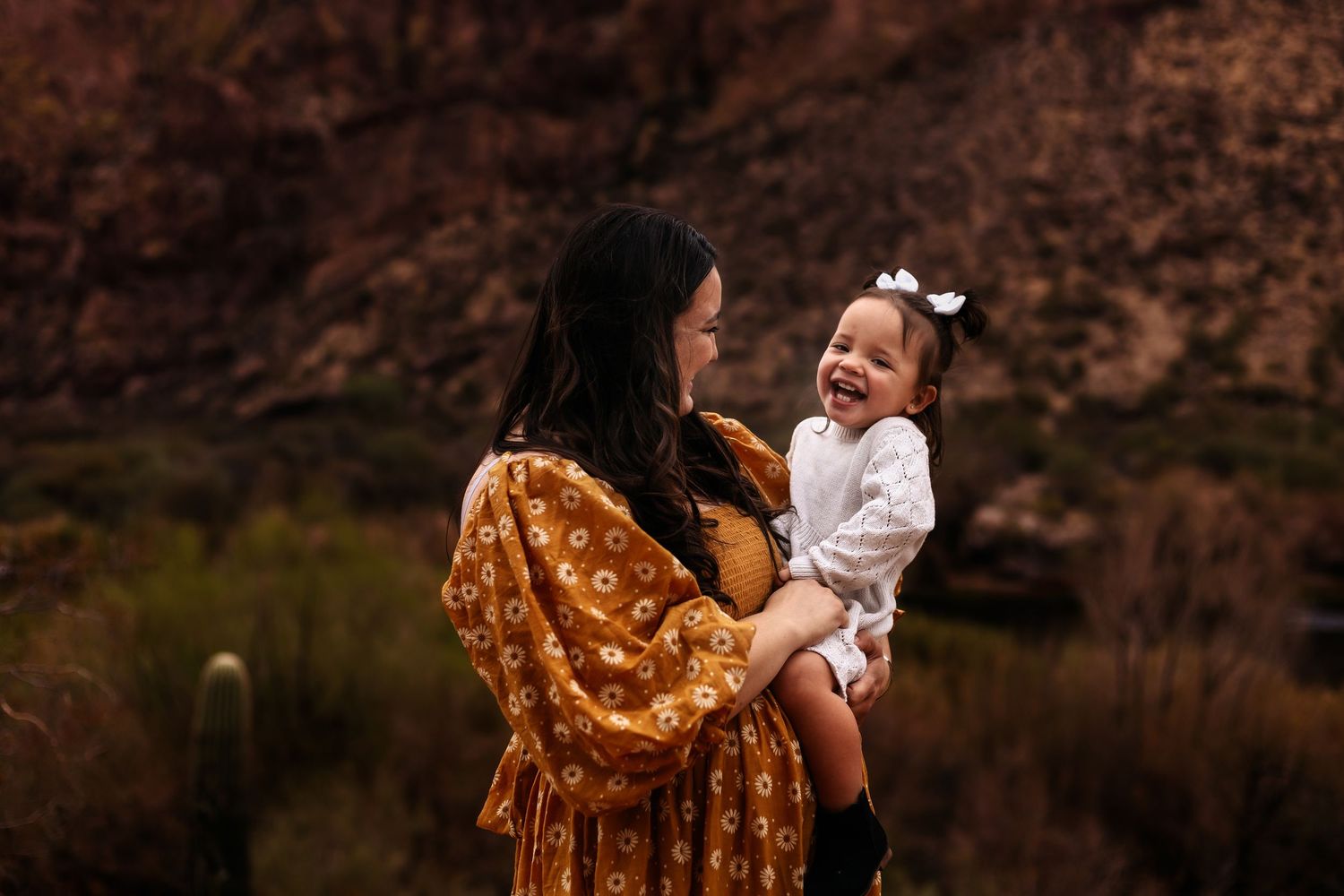 A figure in a mustard floral dress holds a small child wearing white against a scenic mountain backdrop at sunset.