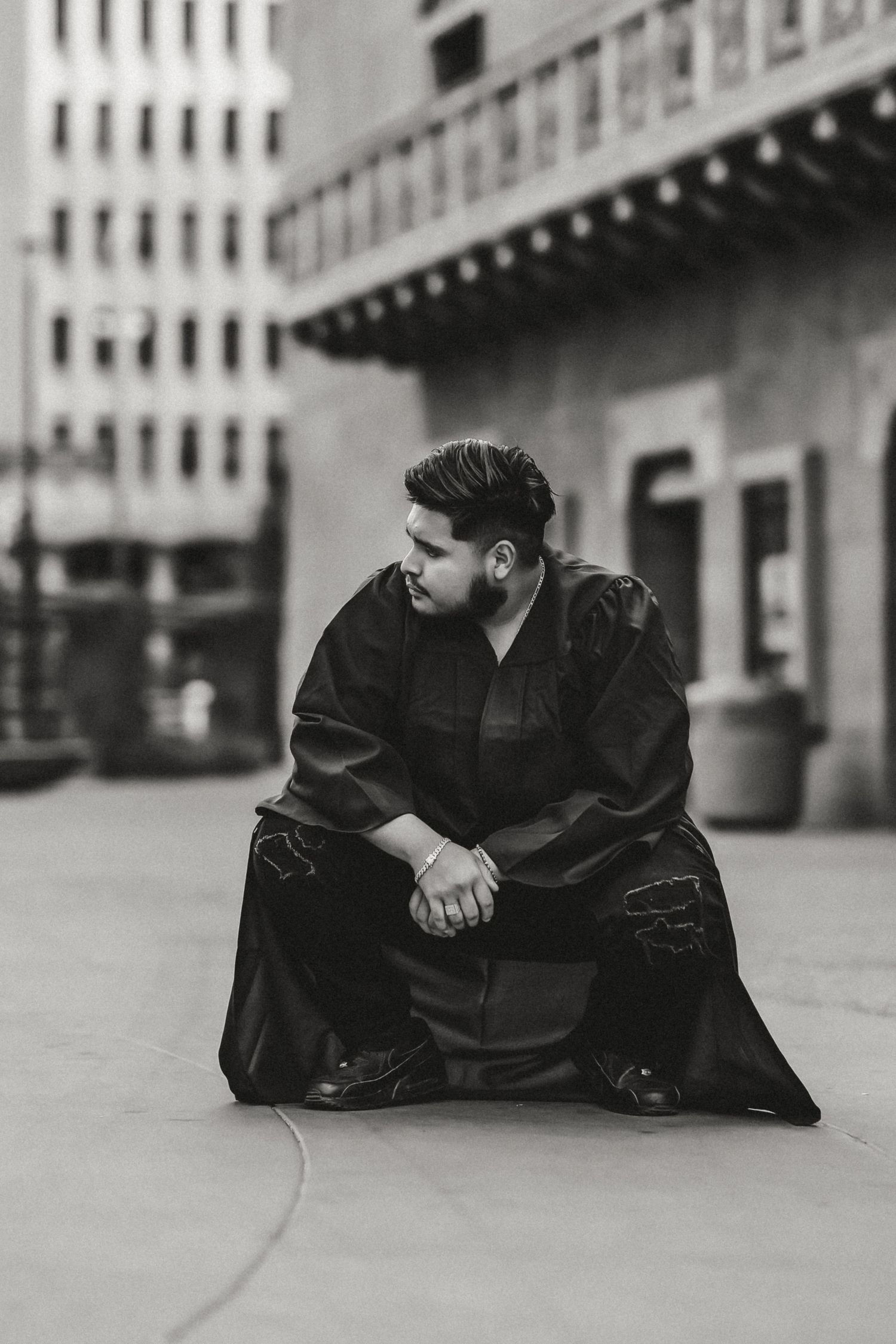 Person in graduation gown sits pensively on urban sidewalk in black and white portrait.