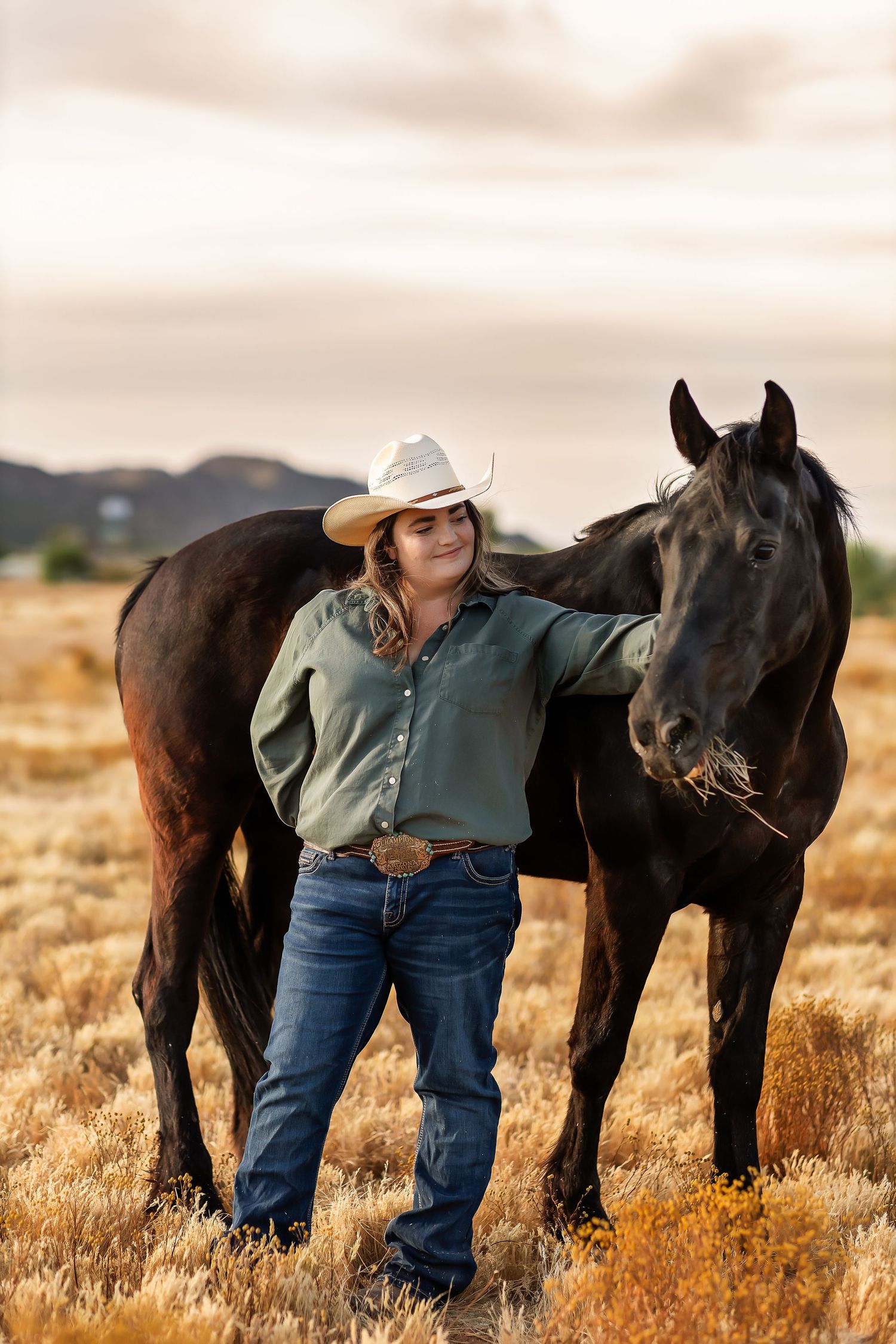 A person in western attire and cowboy hat stands beside a black horse in a golden field at sunset.