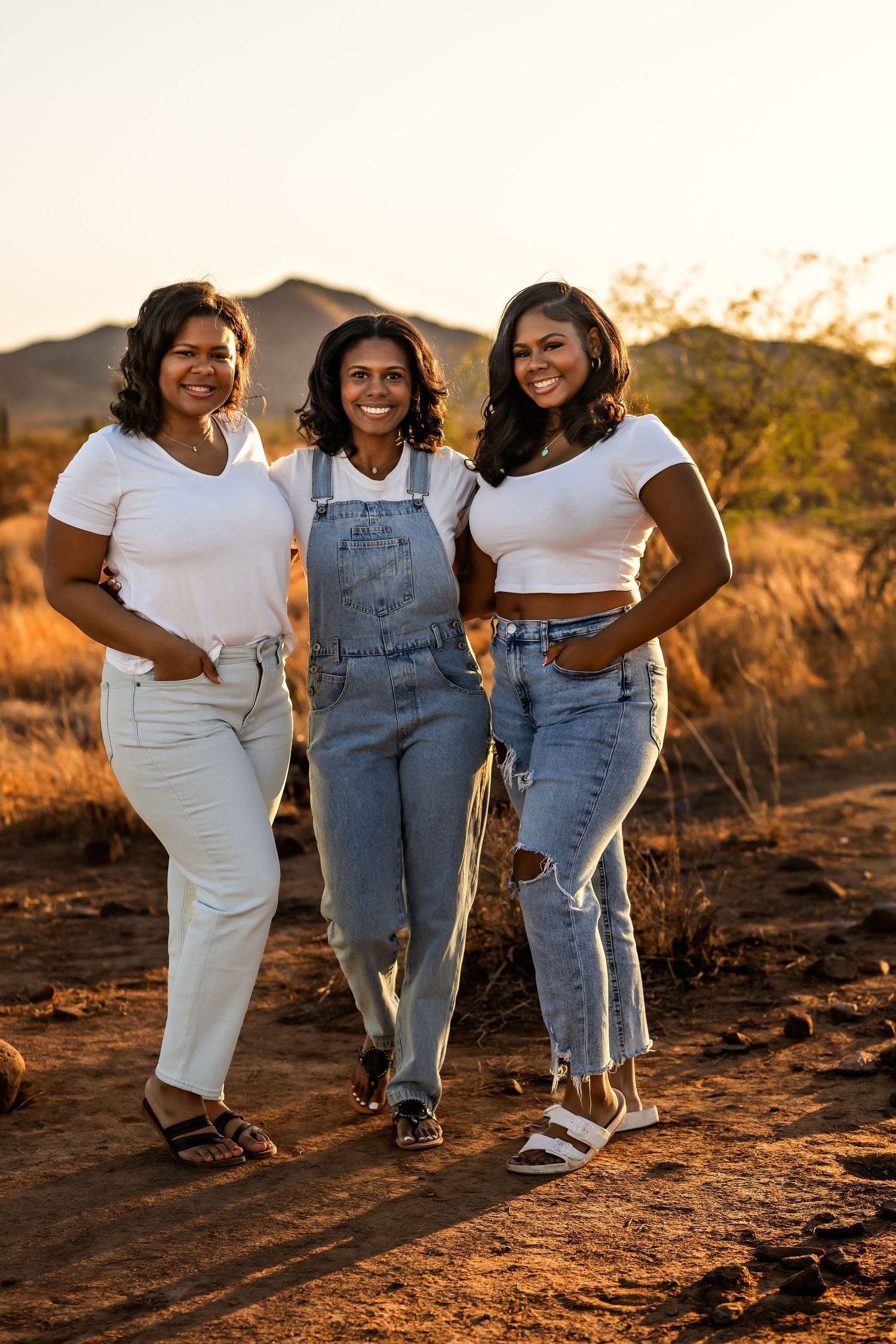 Three friends pose together during golden hour in casual outfits against a desert mountain backdrop.