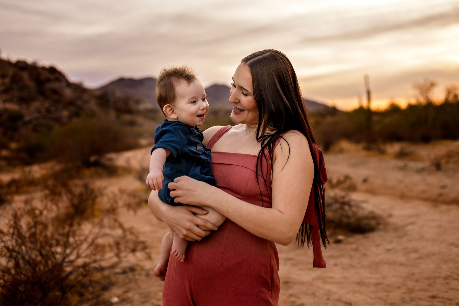 A mother holds a small child in a desert setting during golden hour with mountains in the background.