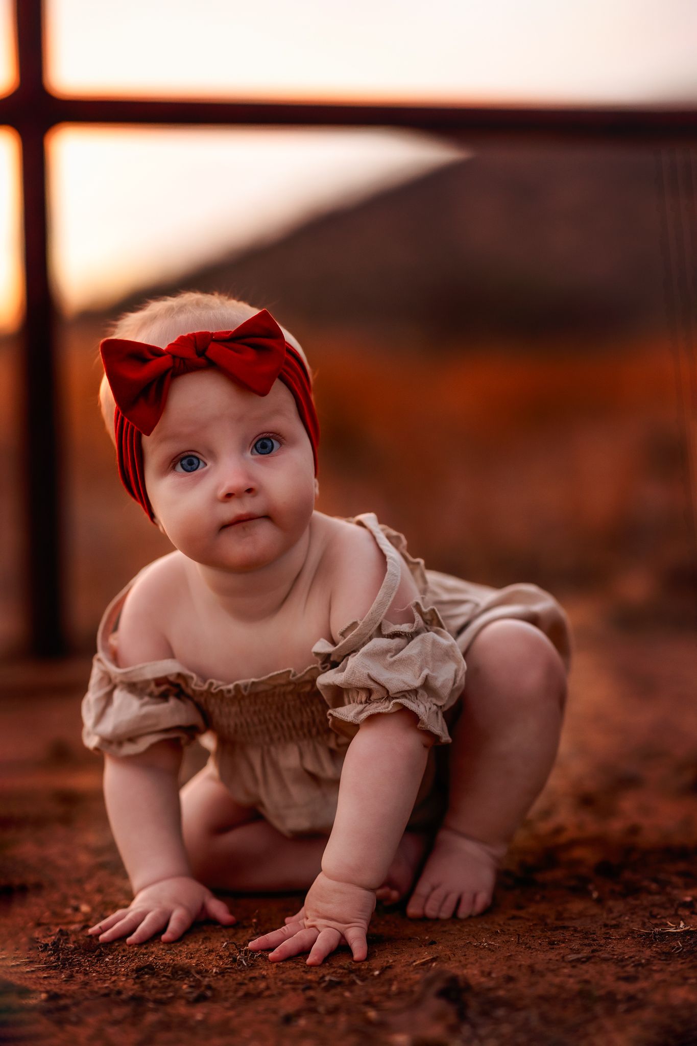 Baby wearing red bow headband and beige ruffled outfit crawls on ground at sunset in warm lighting.