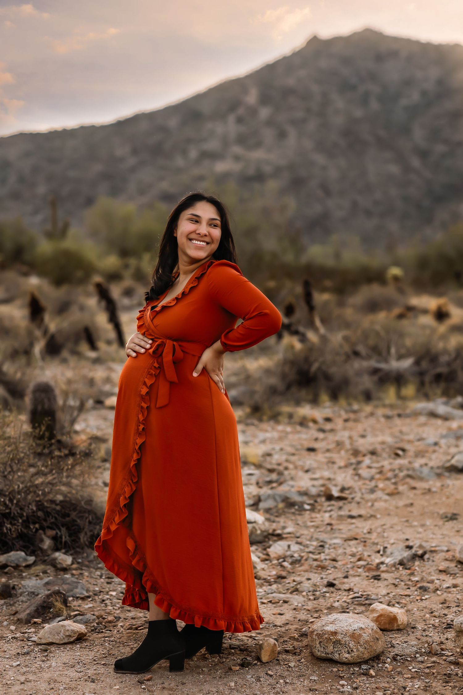 A rust-colored dress flows in the desert breeze during a maternity photoshoot with mountain backdrop.