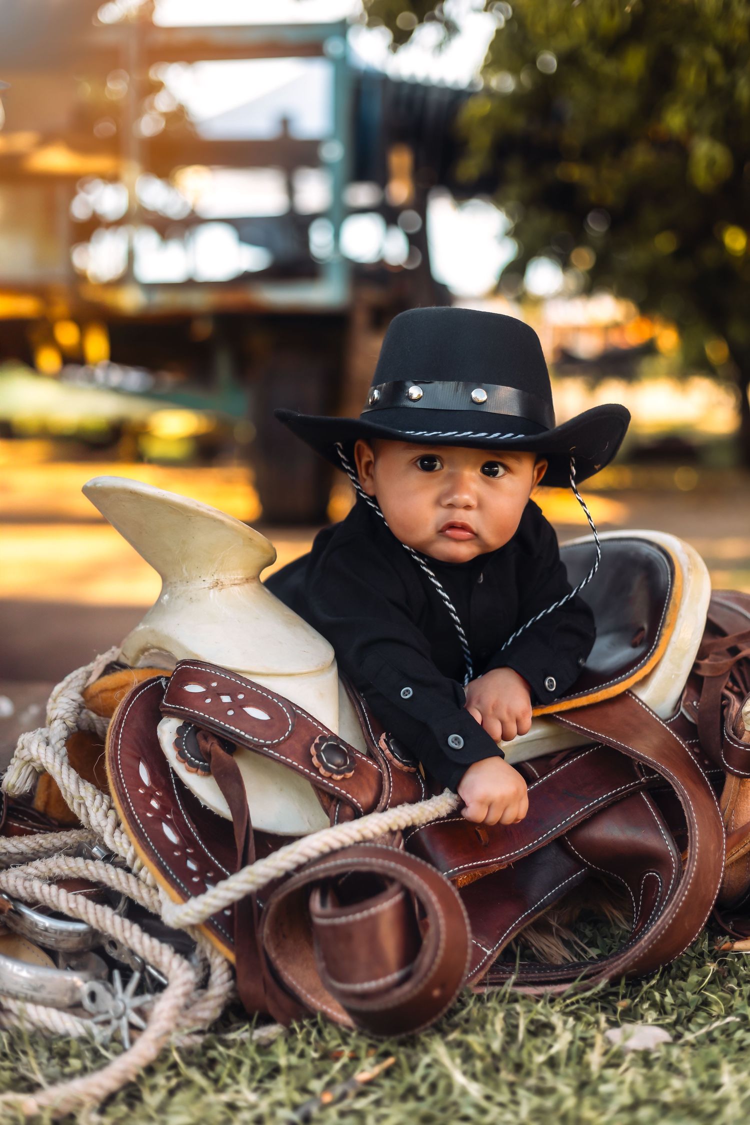 A small child wearing a black cowboy hat sits in a western saddle against a warm sunset background for his milestone photoshoot.