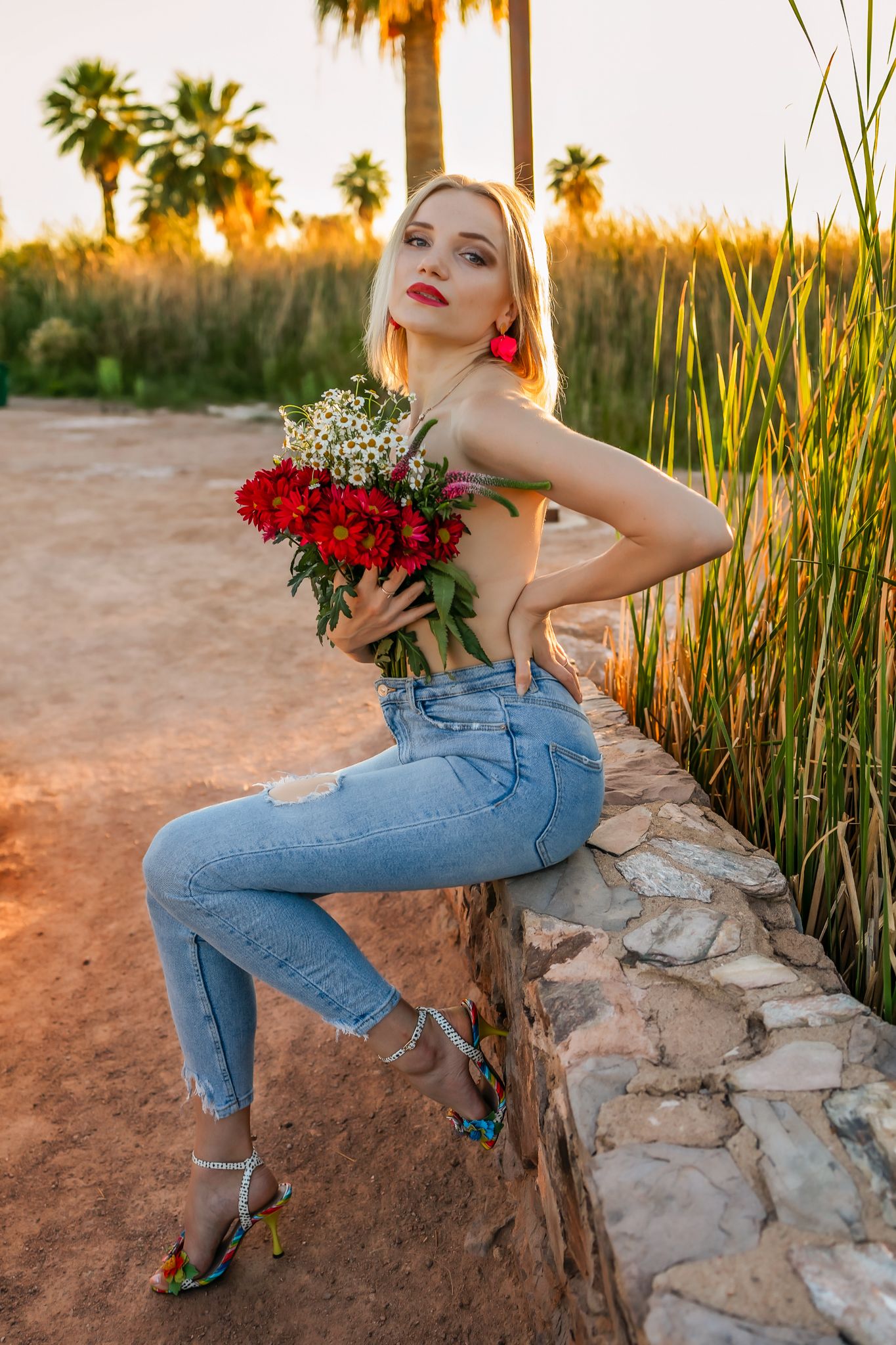 Woman in jeans poses with red roses against palm trees in golden sunset light.
