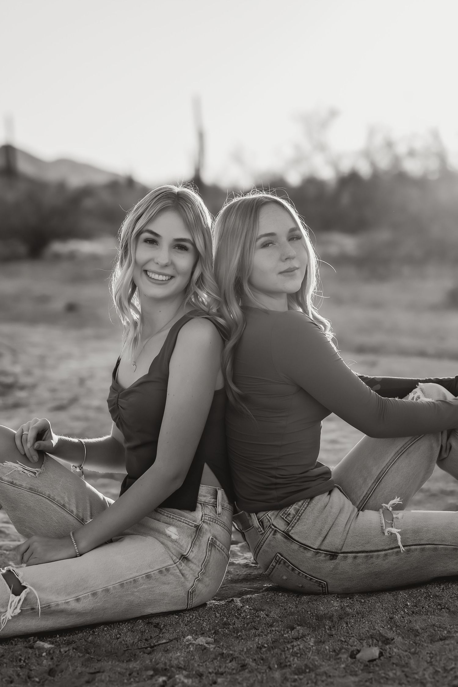 Two sisters in casual wear sitting back-to-back outdoors during sunset for their family photo session.
