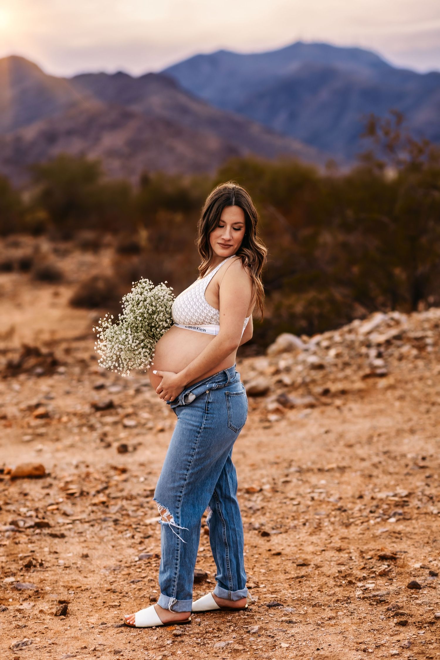 A pregnant woman in jeans holds baby's breath flowers against a desert mountain backdrop.