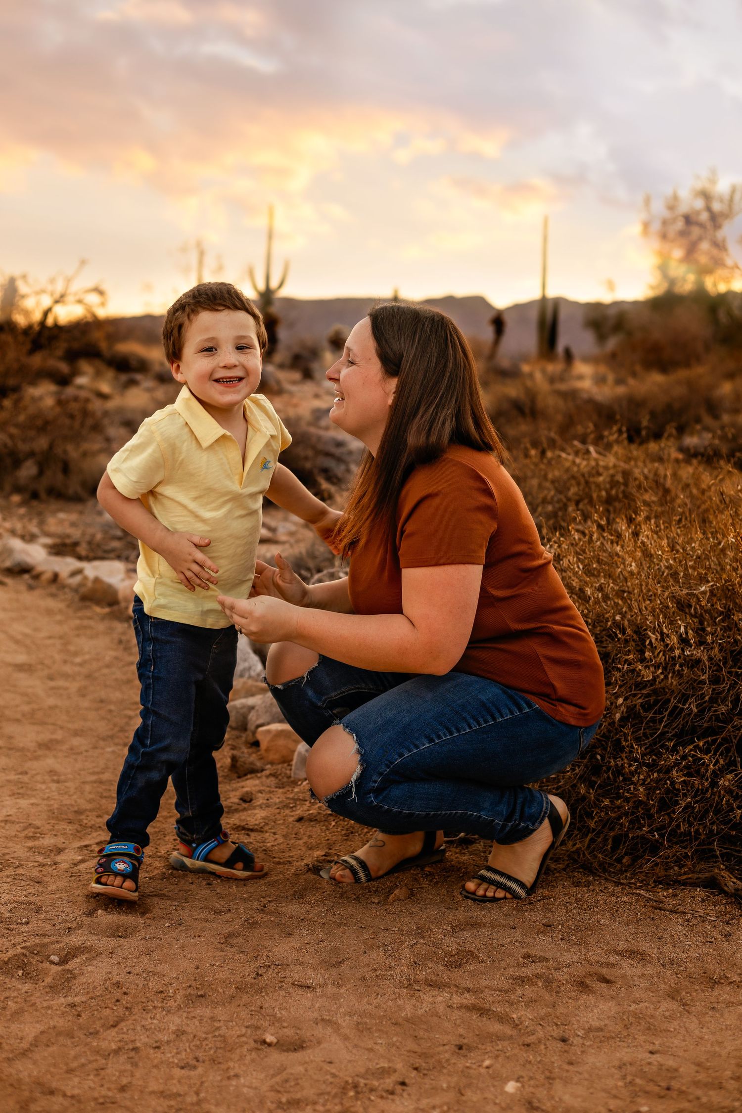 A parent kneels beside a young child on a desert trail during golden hour with cacti in background.