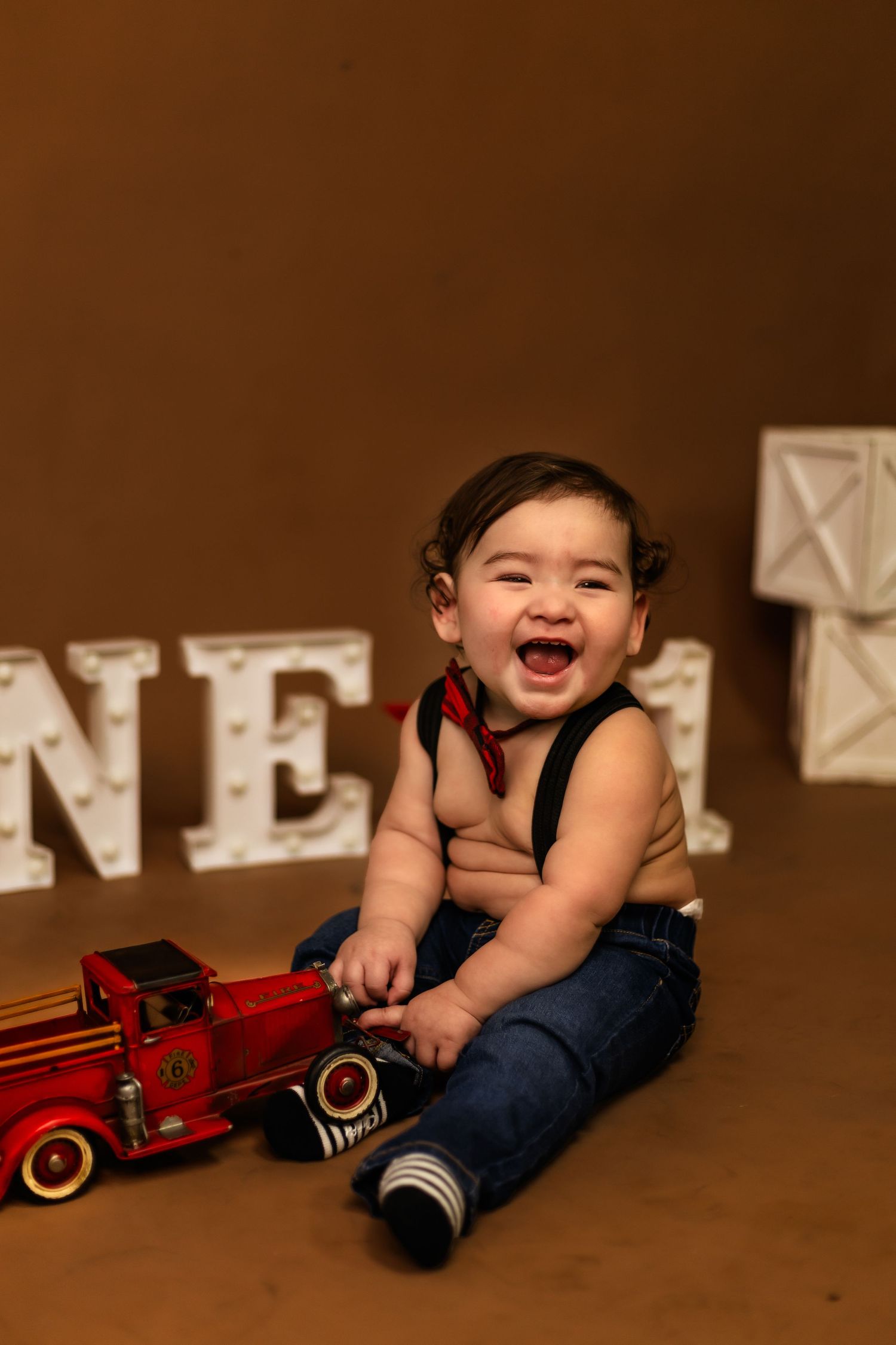 A laughing baby sits next to illuminated ONE letters and a red toy truck against a brown background for his first year milestone photoshoot.