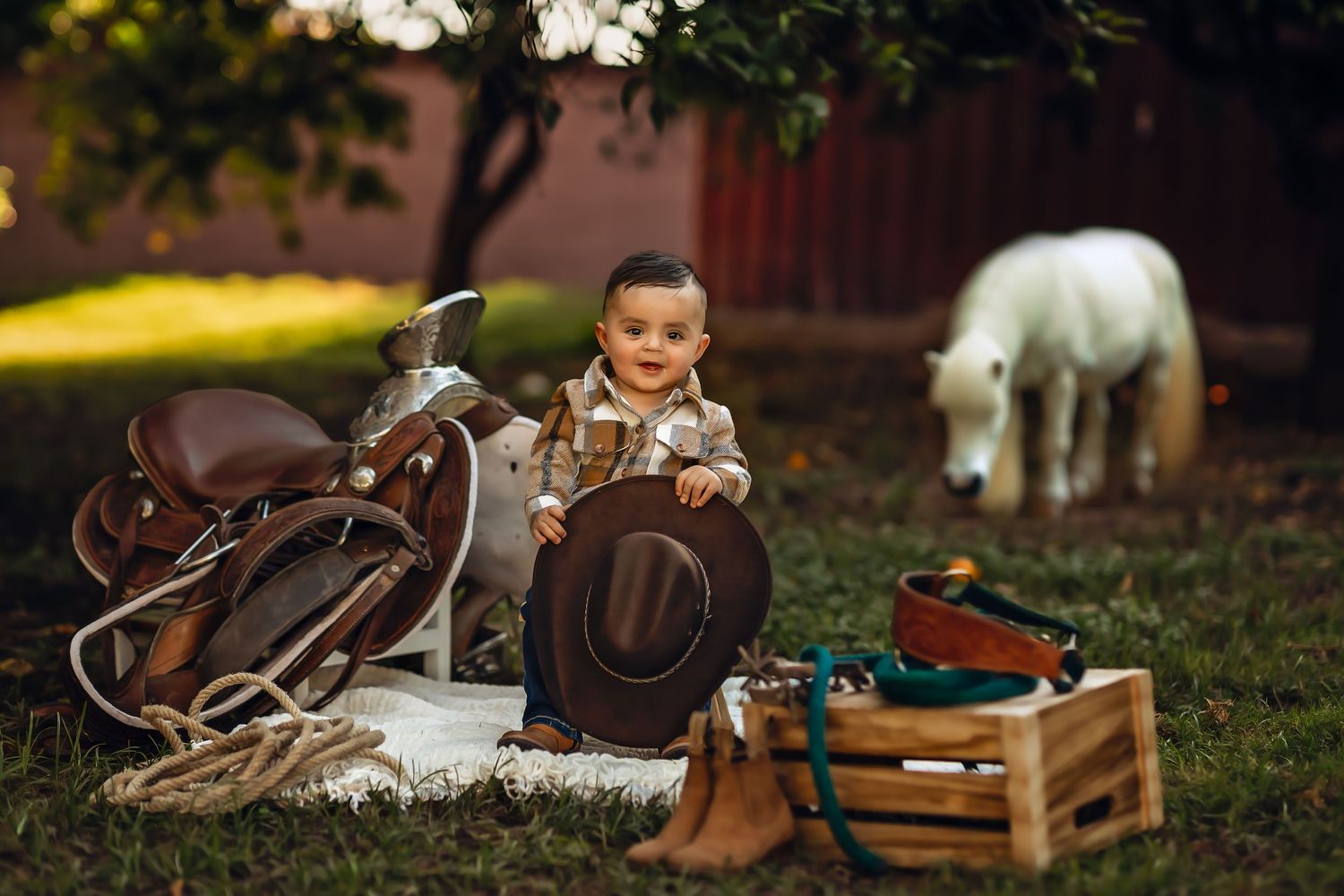 A small child sits among western riding gear and a saddle with a white horse grazing in the background for his milestone photoshoot.