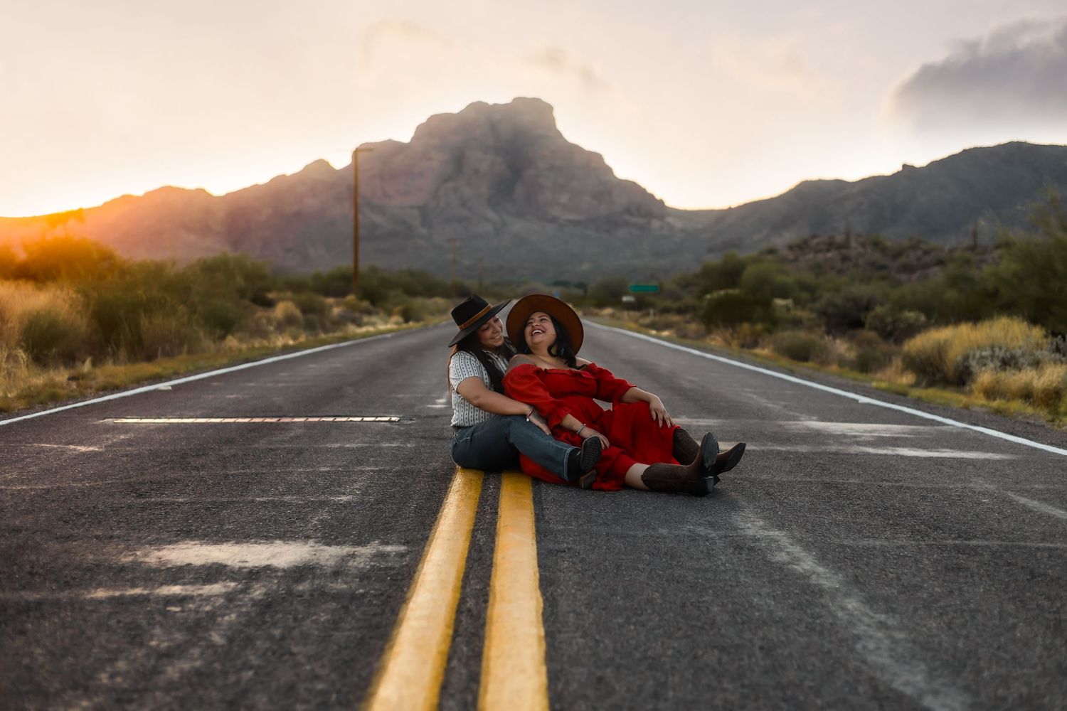 Two people in red lying on yellow lines of empty desert road with mountain backdrop at sunset.