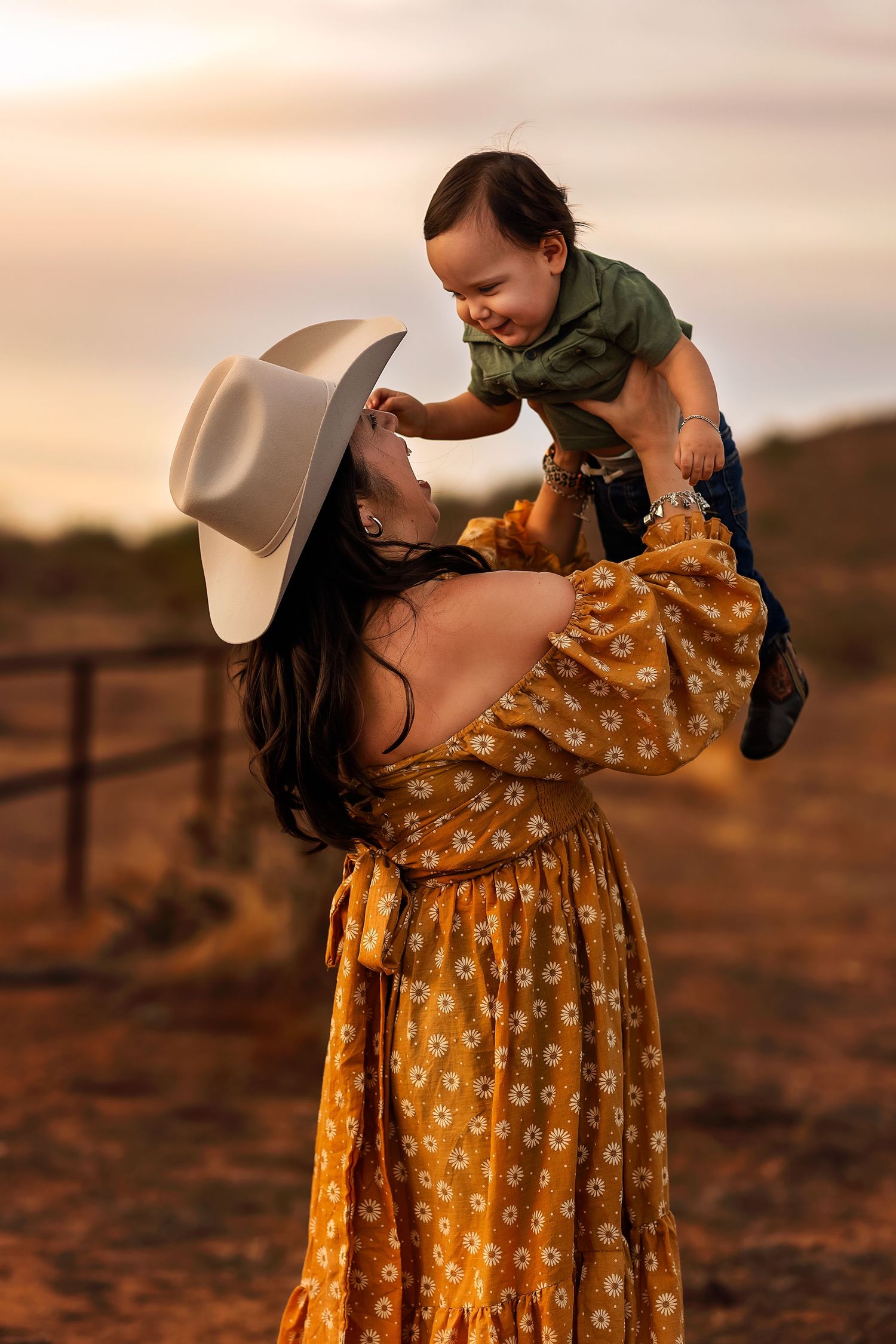 A woman in a floral dress lifts a baby wearing green against a sunset backdrop.