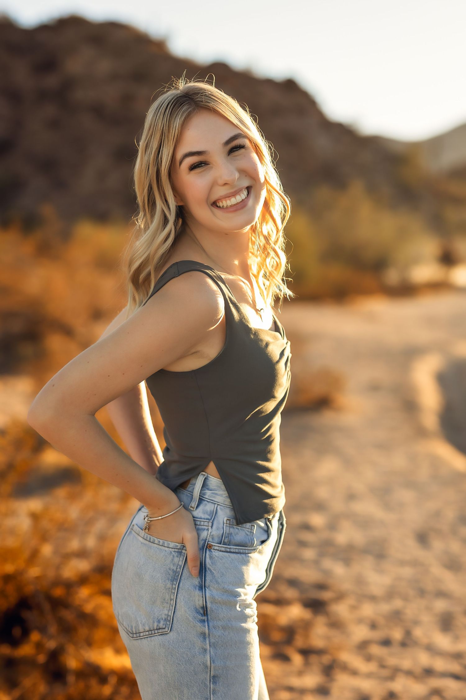 A casual portrait of someone in a tank top and jeans posing against a desert landscape at golden hour.