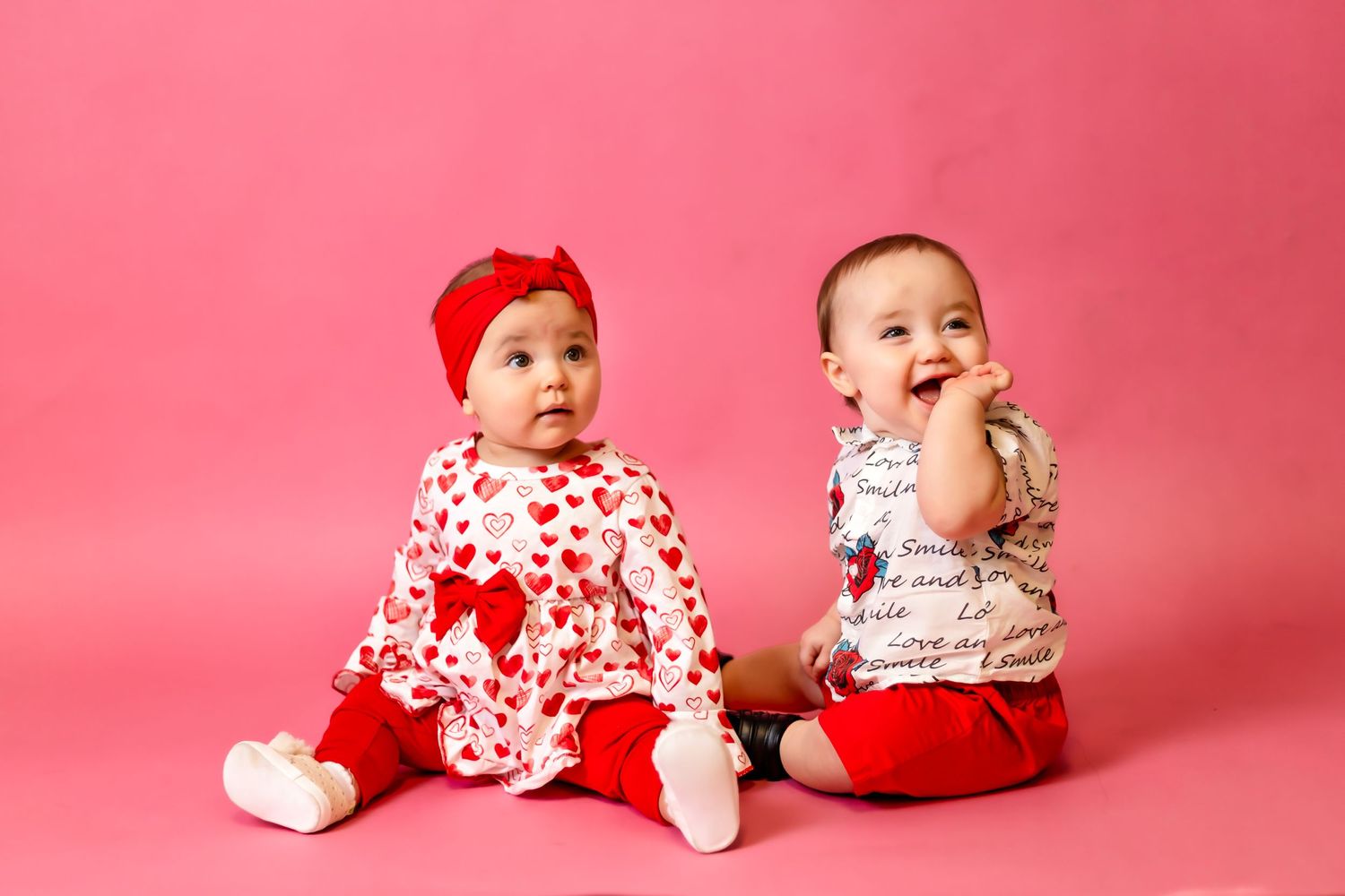 Two babies in red and white outfits laughing against pink backdrop.