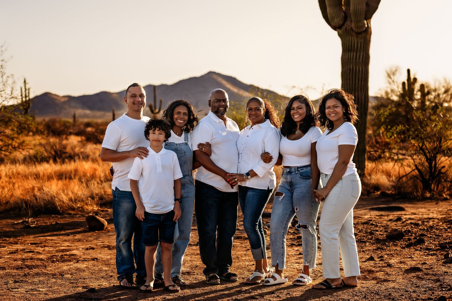 A happy group of people in white shirts and casual clothing pose together on a desert hillside at sunset.