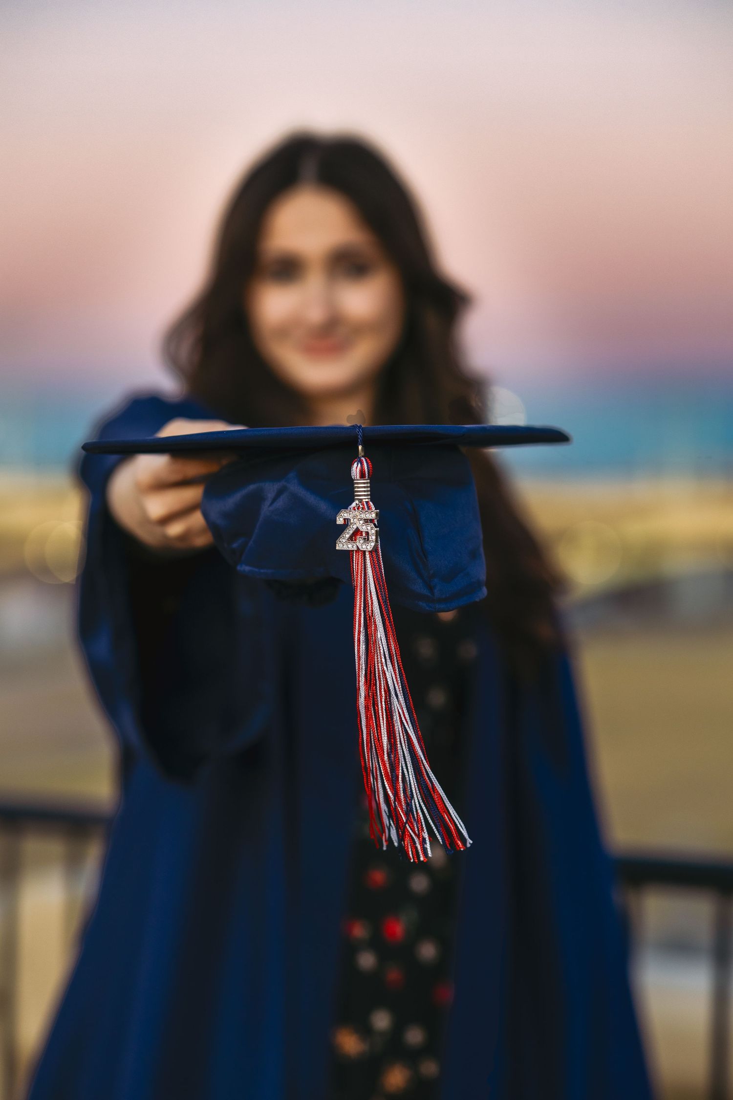 Graduate holding cap with tassel against sunset sky in blue graduation gown in Tempe Arizona.