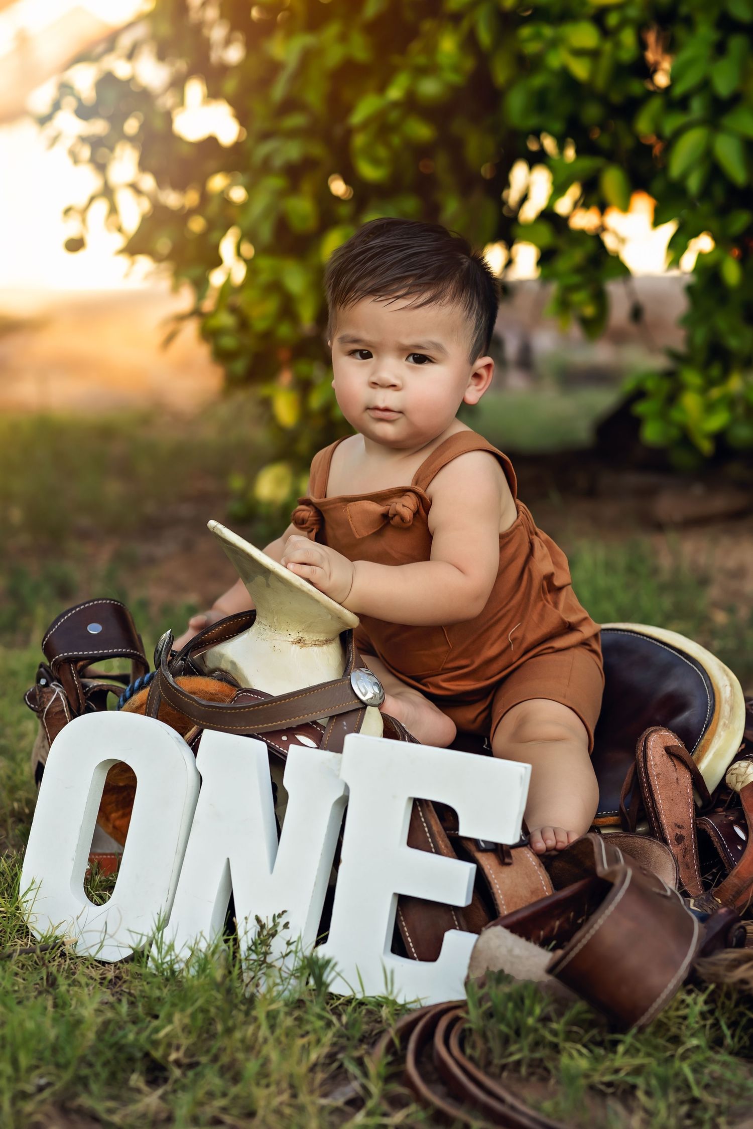 A one-year-old in brown overalls sits with white ONE letters and cowboy boots in an outdoor setting for his first birthday cake smash.