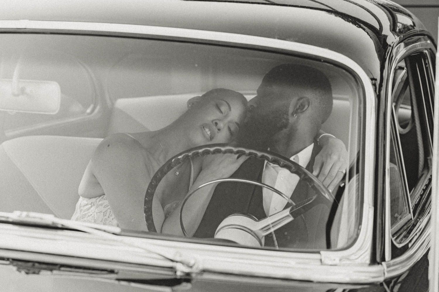 A couple sits close together in a vintage car while one person rests their head on the other's shoulder.