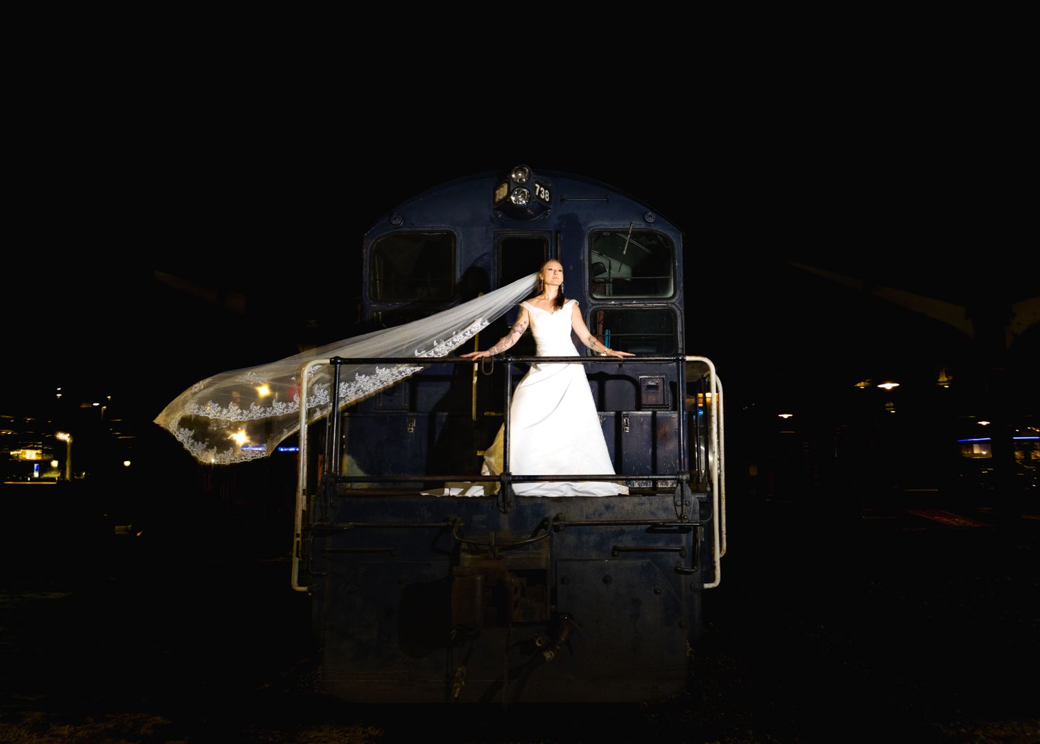 A person in a white wedding dress stands illuminated on a dark train platform at night.