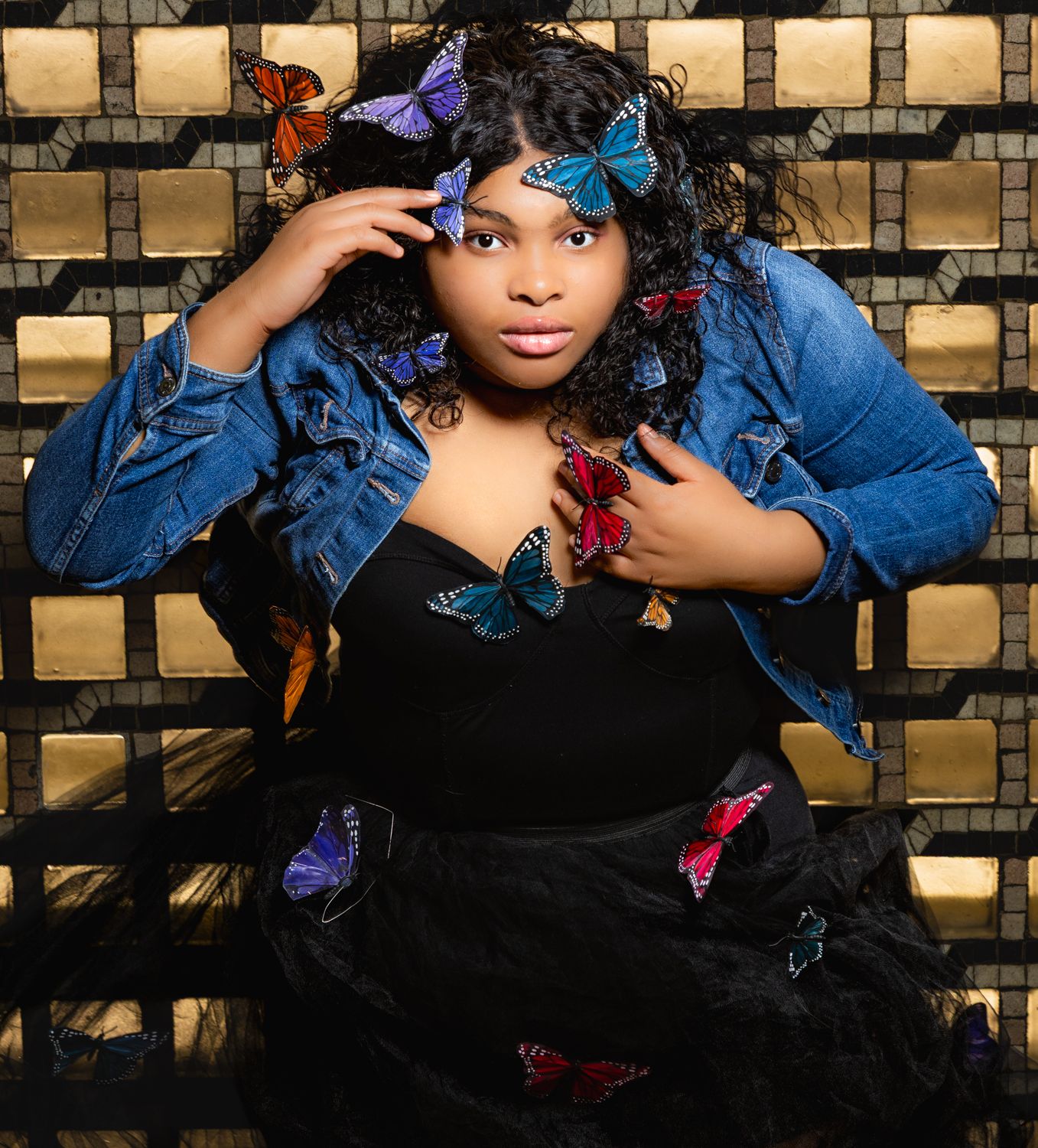 Multiple butterflies surround a person wearing a black dress and denim jacket in a dark atmospheric portrait session.