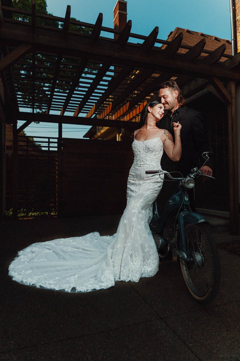 A couple sharing an intimate moment under a wooden pergola with a bicycle nearby during a sunset wedding photoshoot.