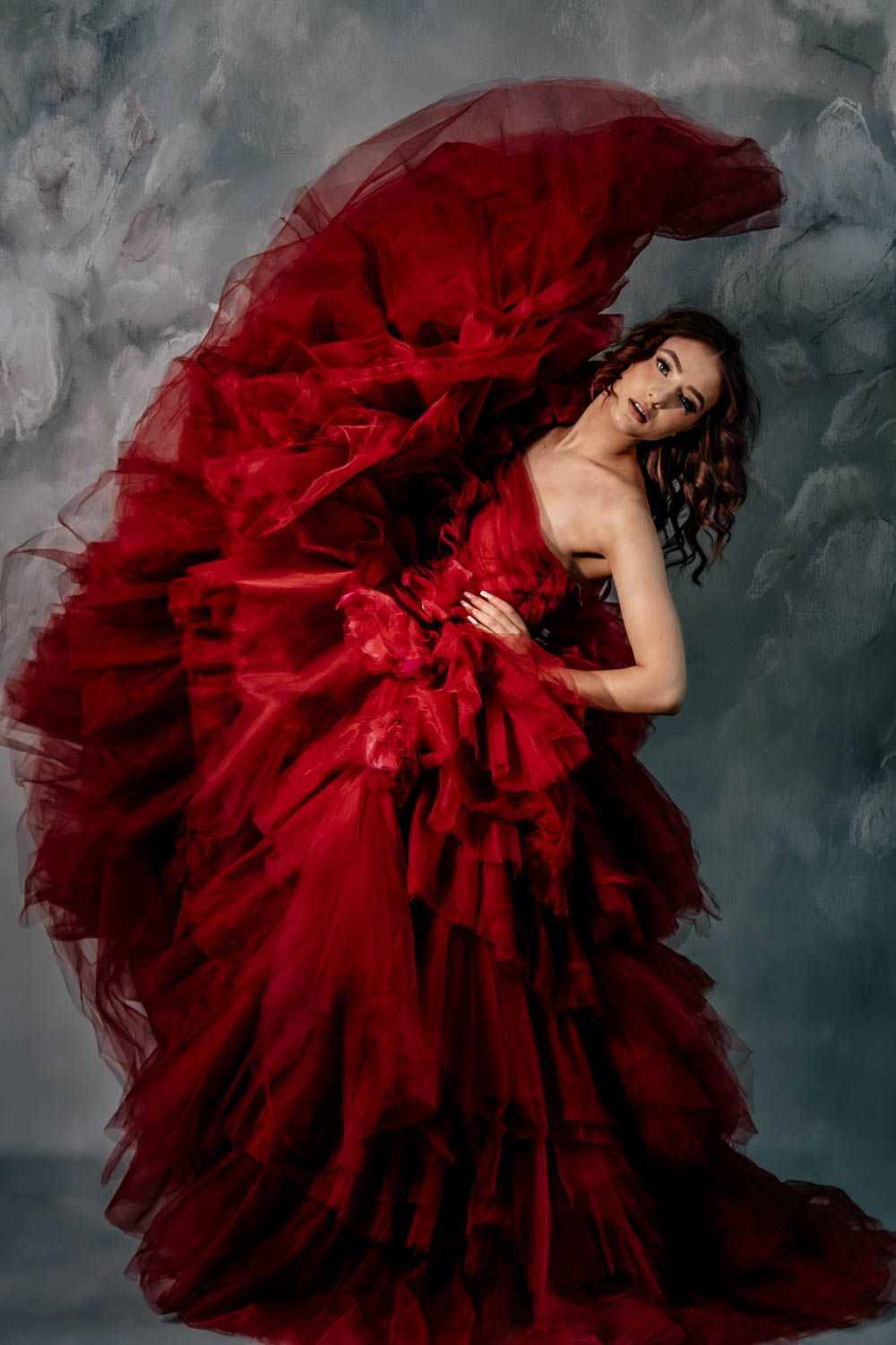 A figure in a dramatic flowing deep red tulle gown poses gracefully against a moody gray backdrop with fog effects.