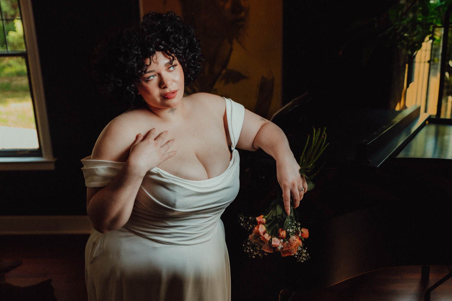 Plus size model in white dress poses near window with moody natural lighting and floral bouquet.