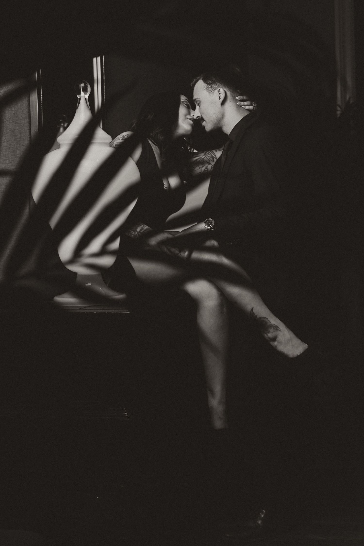 A dramatic black and white photo of a couple embracing in shadows with striking light patterns cast across them.