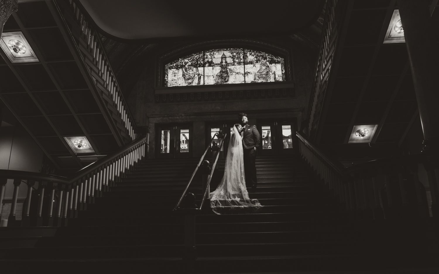 A dramatic black and white photograph of a couple standing on an ornate staircase in a dimly lit elegant venue.
