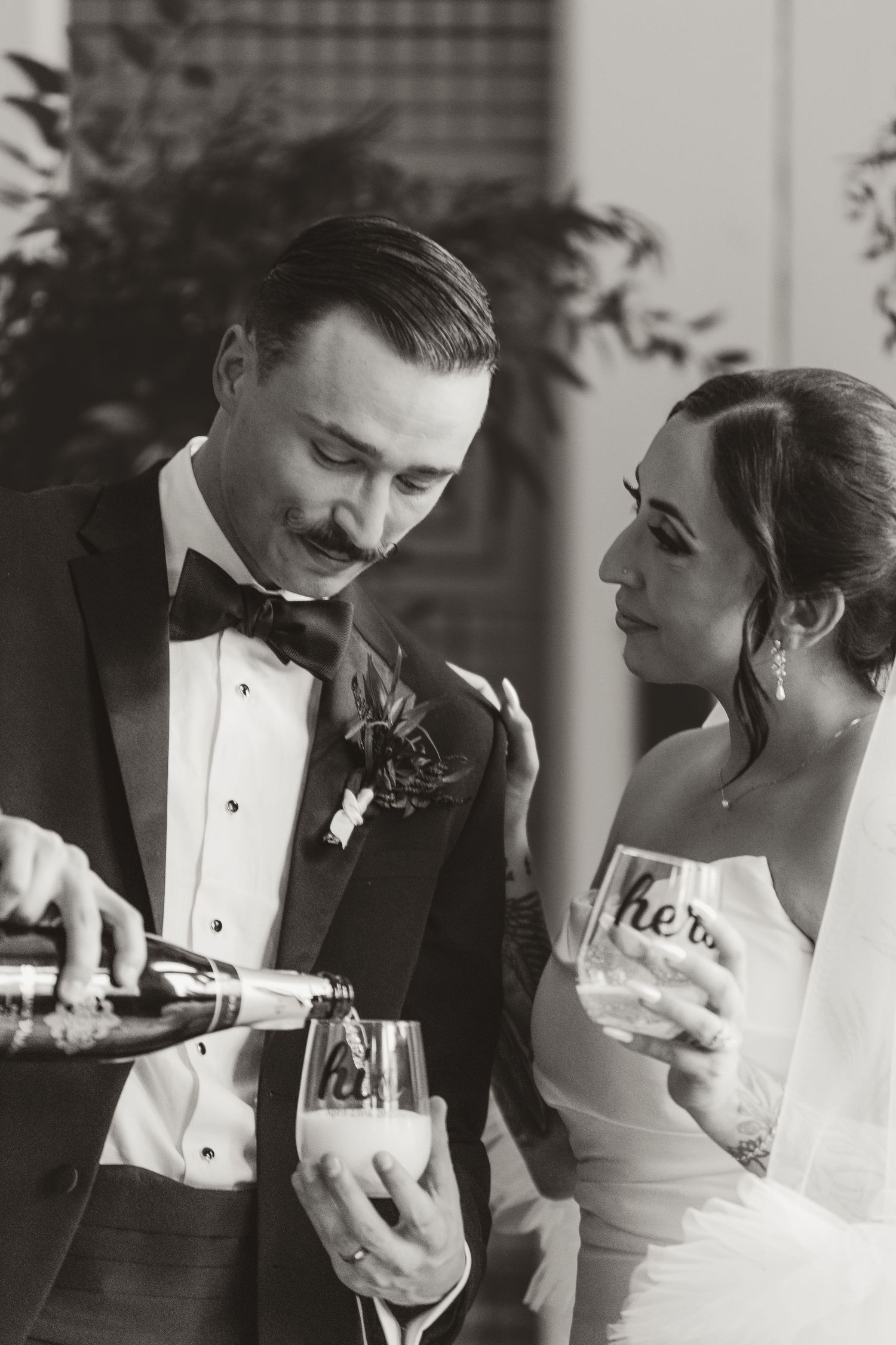 Black and white photo of a bride and groom sharing a champagne moment together in formal wedding attire.