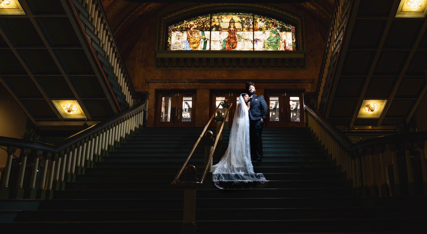 A couple in wedding attire stands on an elegant wooden staircase beneath a stained glass window in dim lighting.