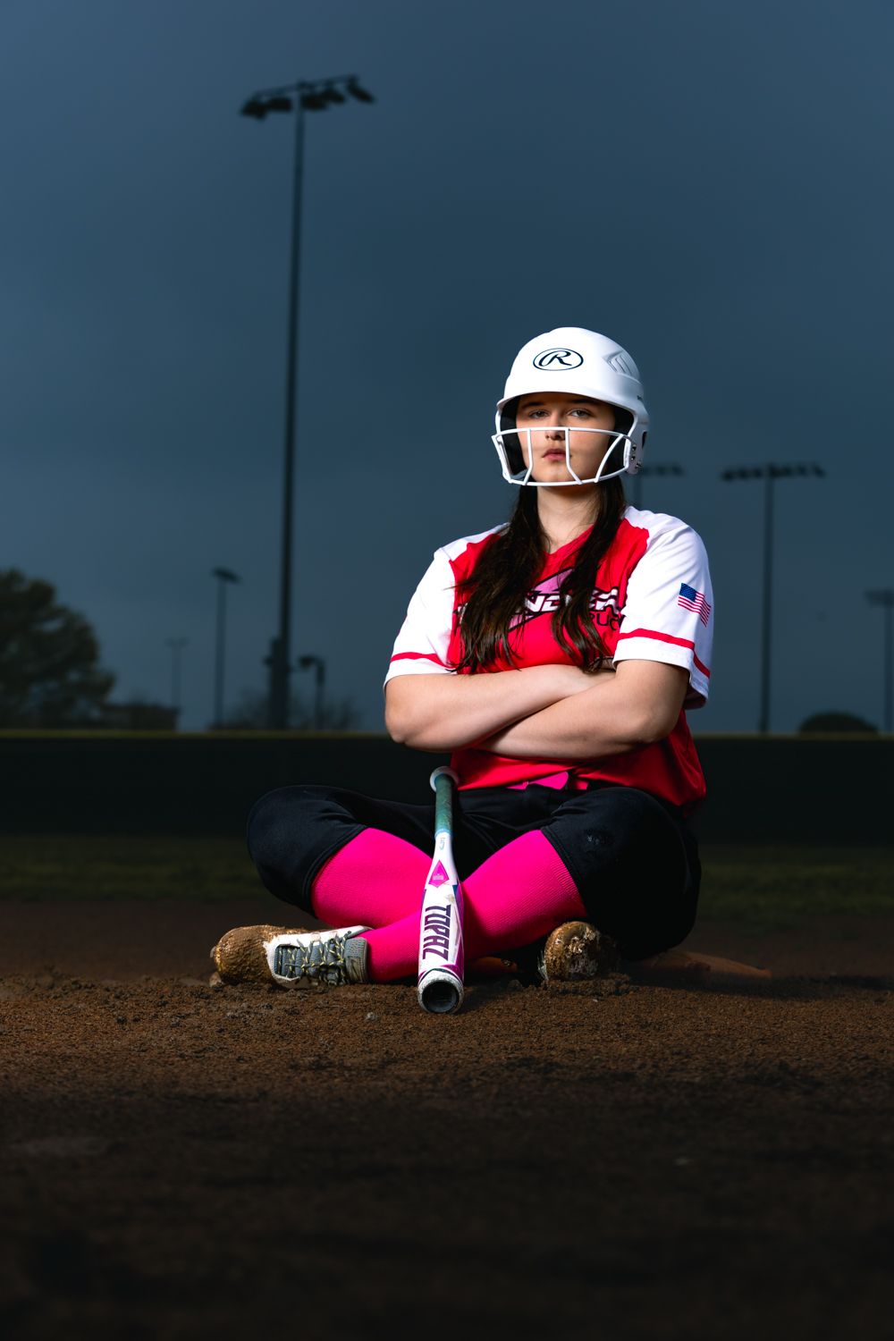 Softball player in white helmet and red jersey sits on field at dusk with pink socks and white bat beside them.