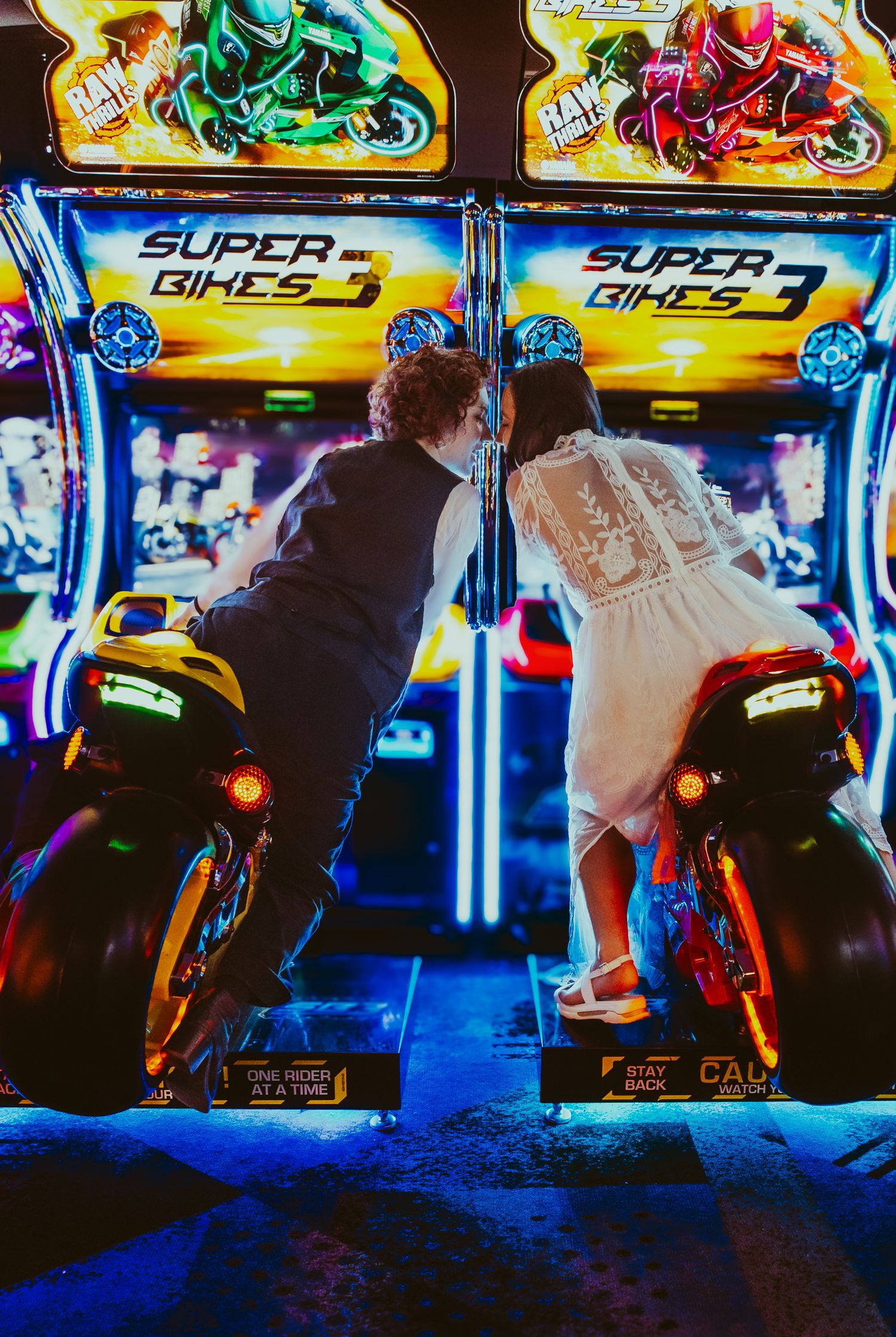 A couple play retro arcade motorcycle racing games together in white and black outfits at a neon-lit arcade.