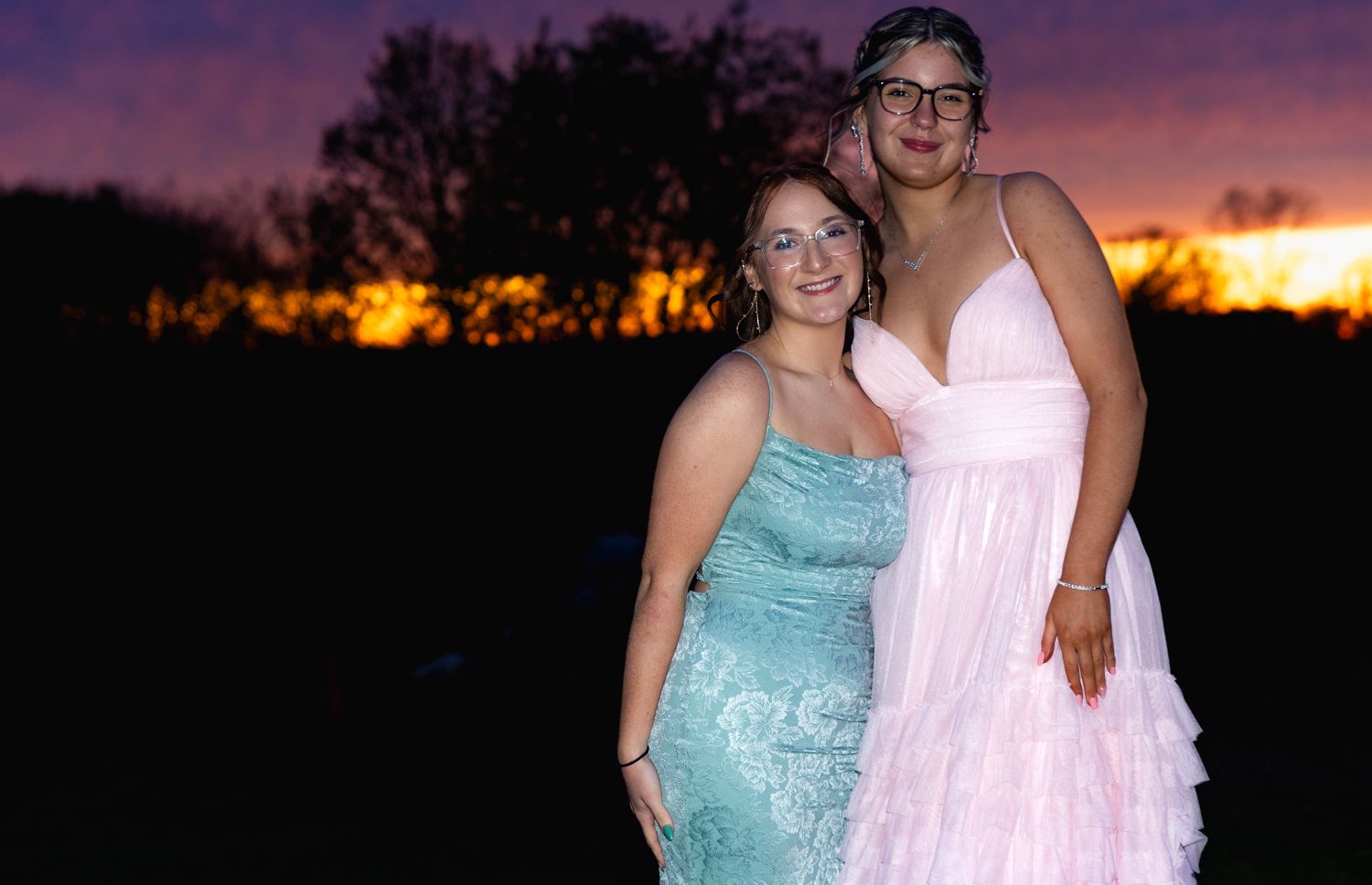 Two prom-goers in formal dresses pose at sunset with a glowing orange and purple sky in the background.