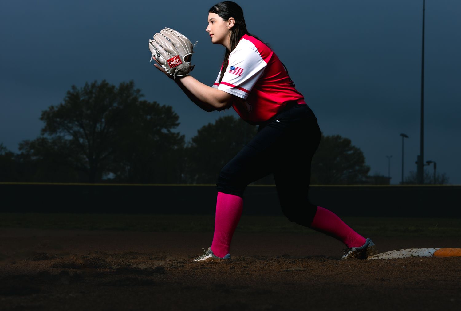 A softball player in a red jersey and black pants fields a ground ball at night on a dirt infield.