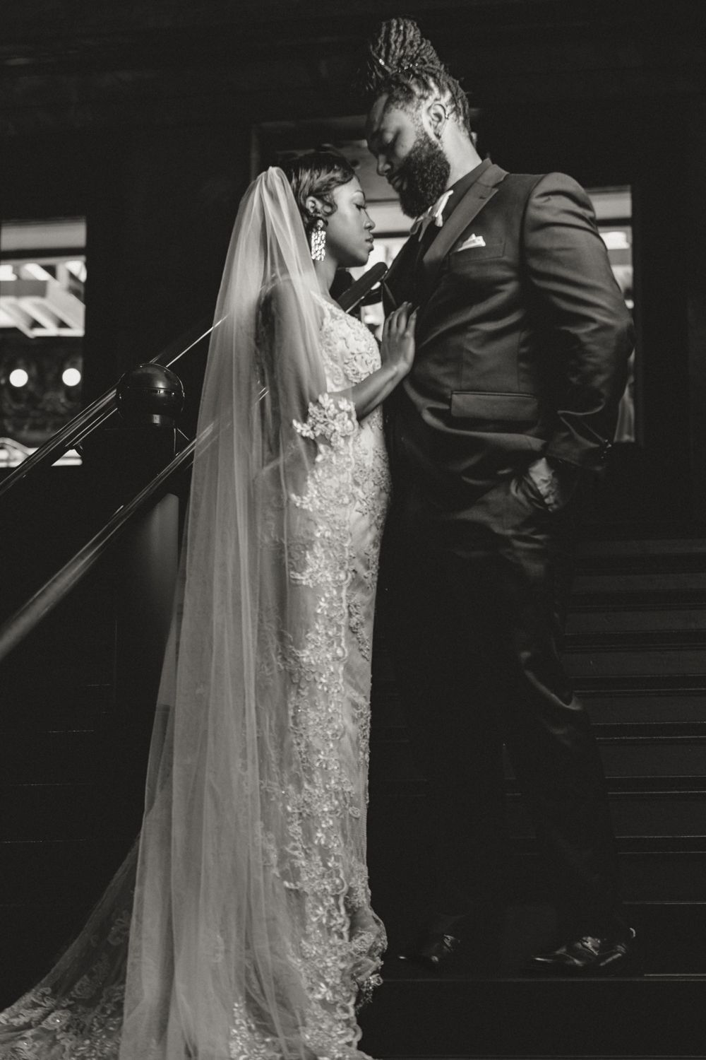 A romantic black and white wedding photo of a bride in lace dress and groom on stairs sharing an intimate moment.
