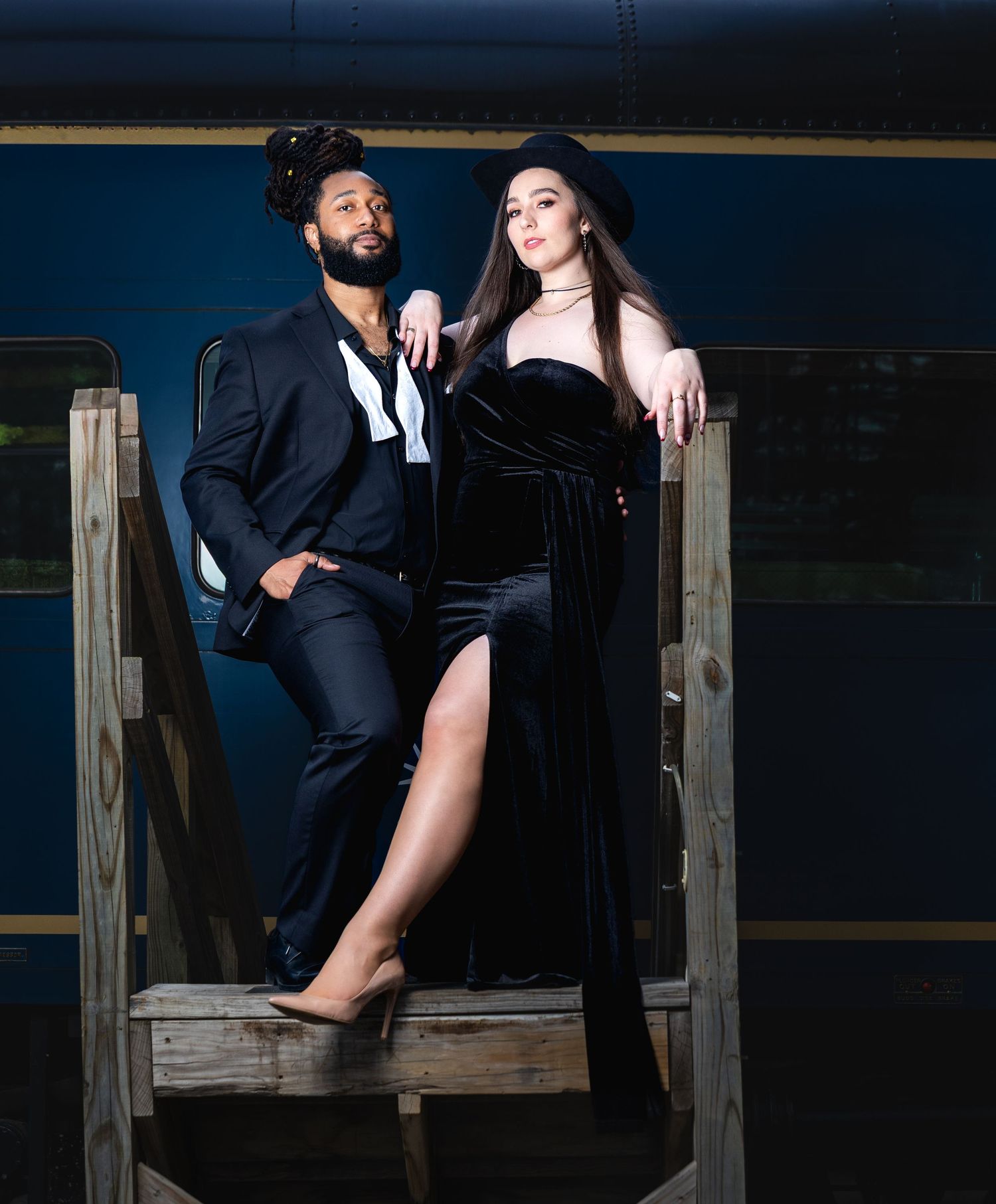 Stylishly dressed couple poses on wooden steps at night wearing elegant black outfits.