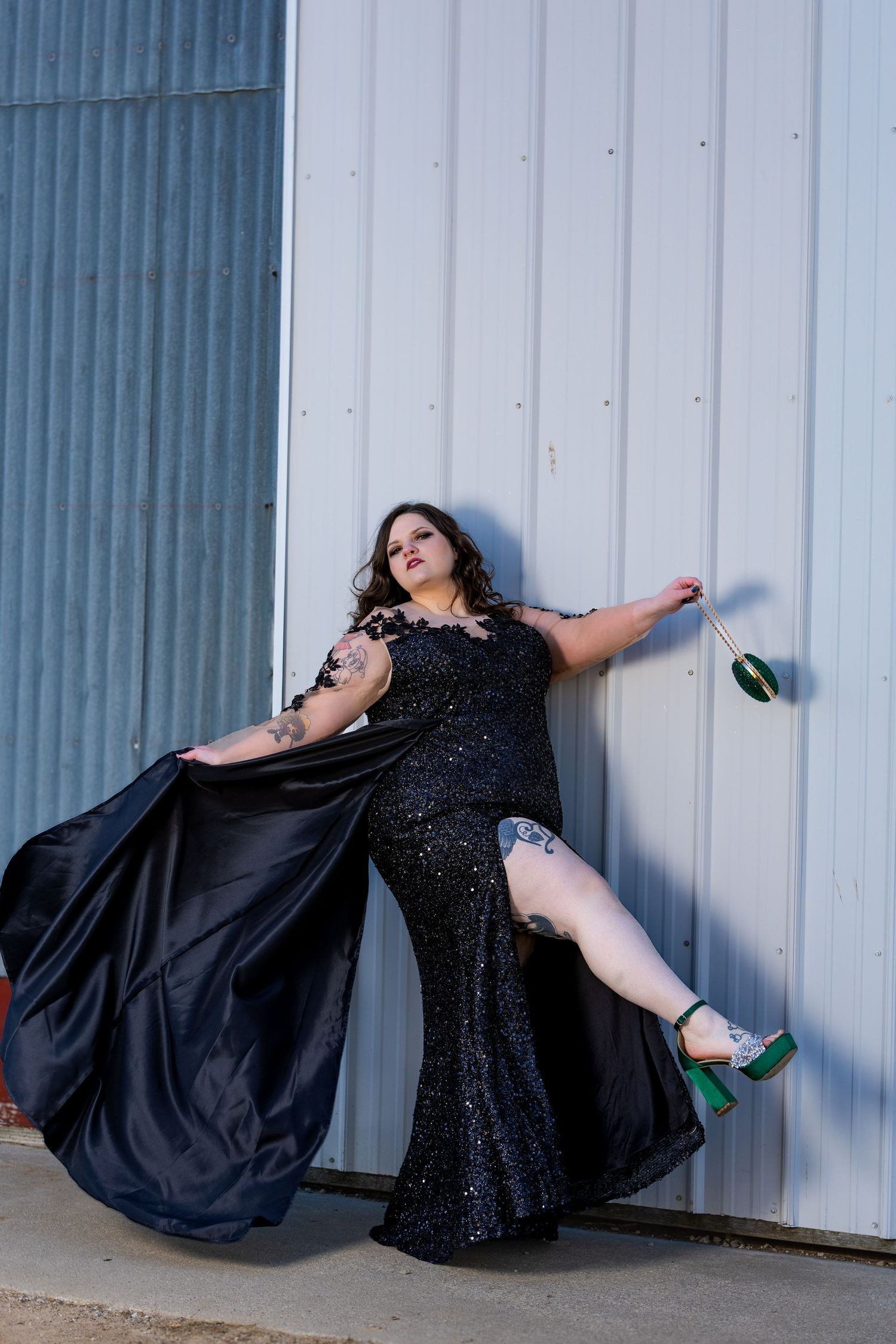 A person in a flowing black sequined evening gown poses dramatically against an industrial metal wall.