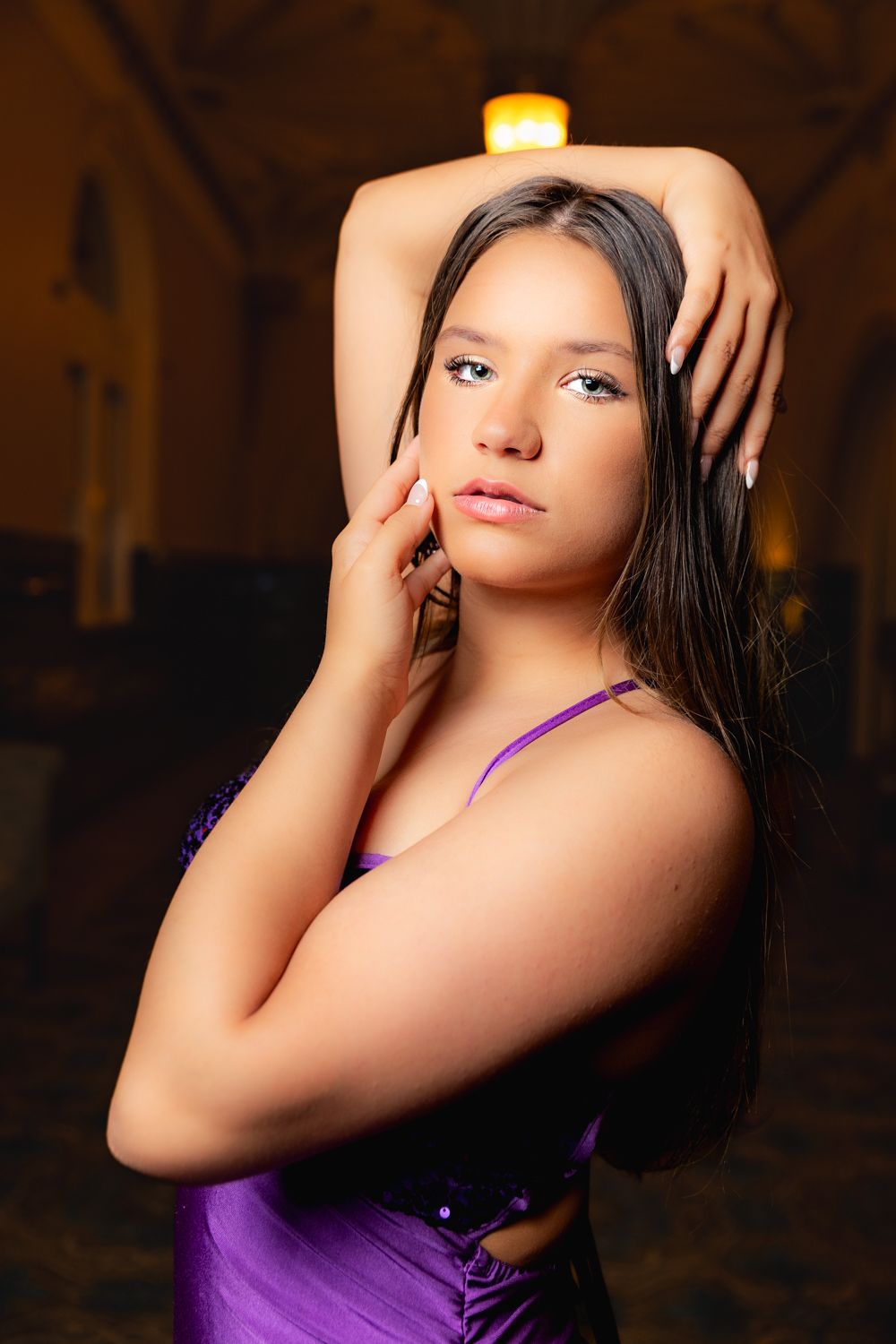 Portrait in moody lighting with dark background features person in purple top striking dramatic pose.