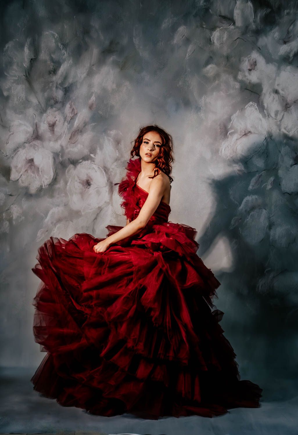 A dramatic portrait features a luxurious red ball gown against a moody grey backdrop with dreamy floral textures.