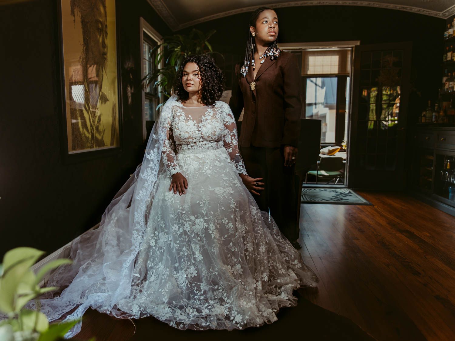Wedding couple pose in an elegant indoor setting with ornate lace gown and dark suit in moody lighting.
