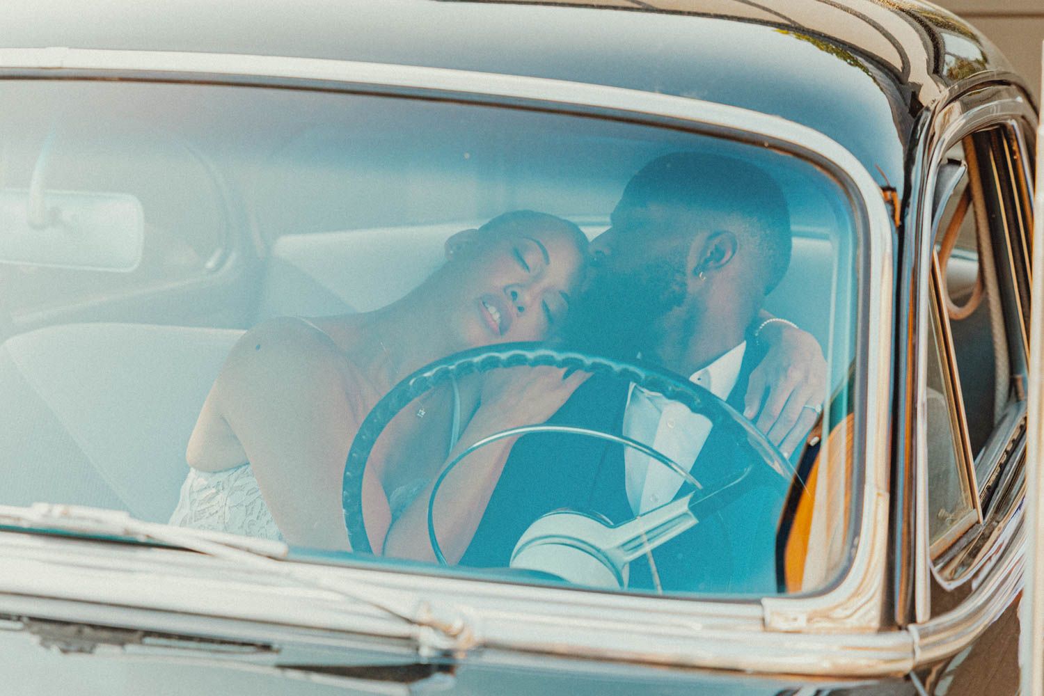A turquoise vintage car with passengers visible through the windshield on a sunny afternoon.