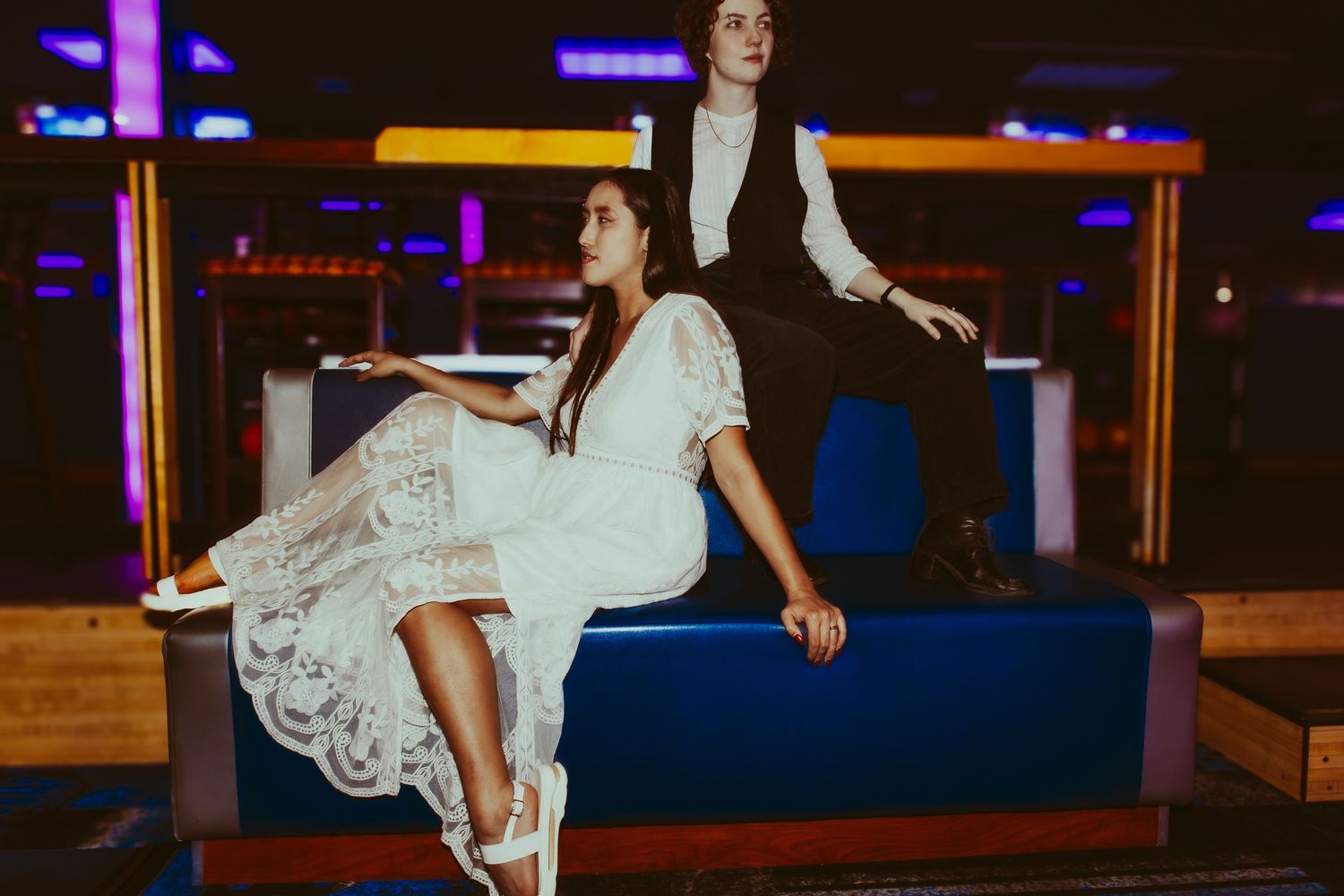 Two people playfully pose on a blue bowling alley bench with colorful neon lights in the background.