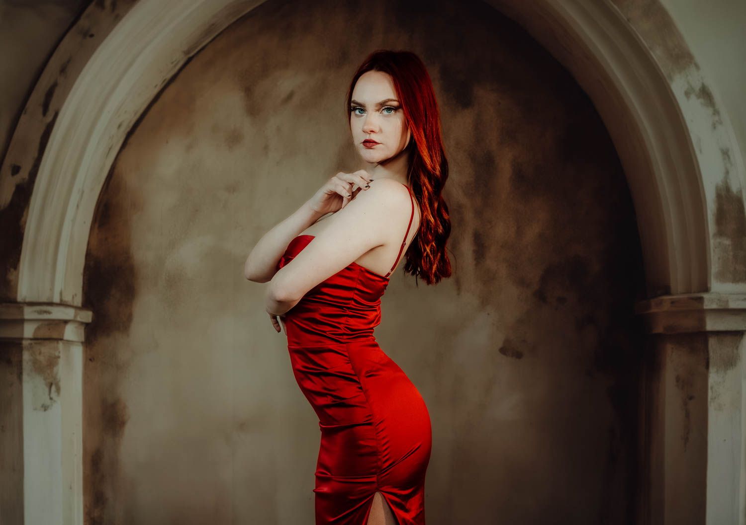Elegant figure in a form-fitting red velvet dress poses in an arched stone doorway with dramatic lighting.